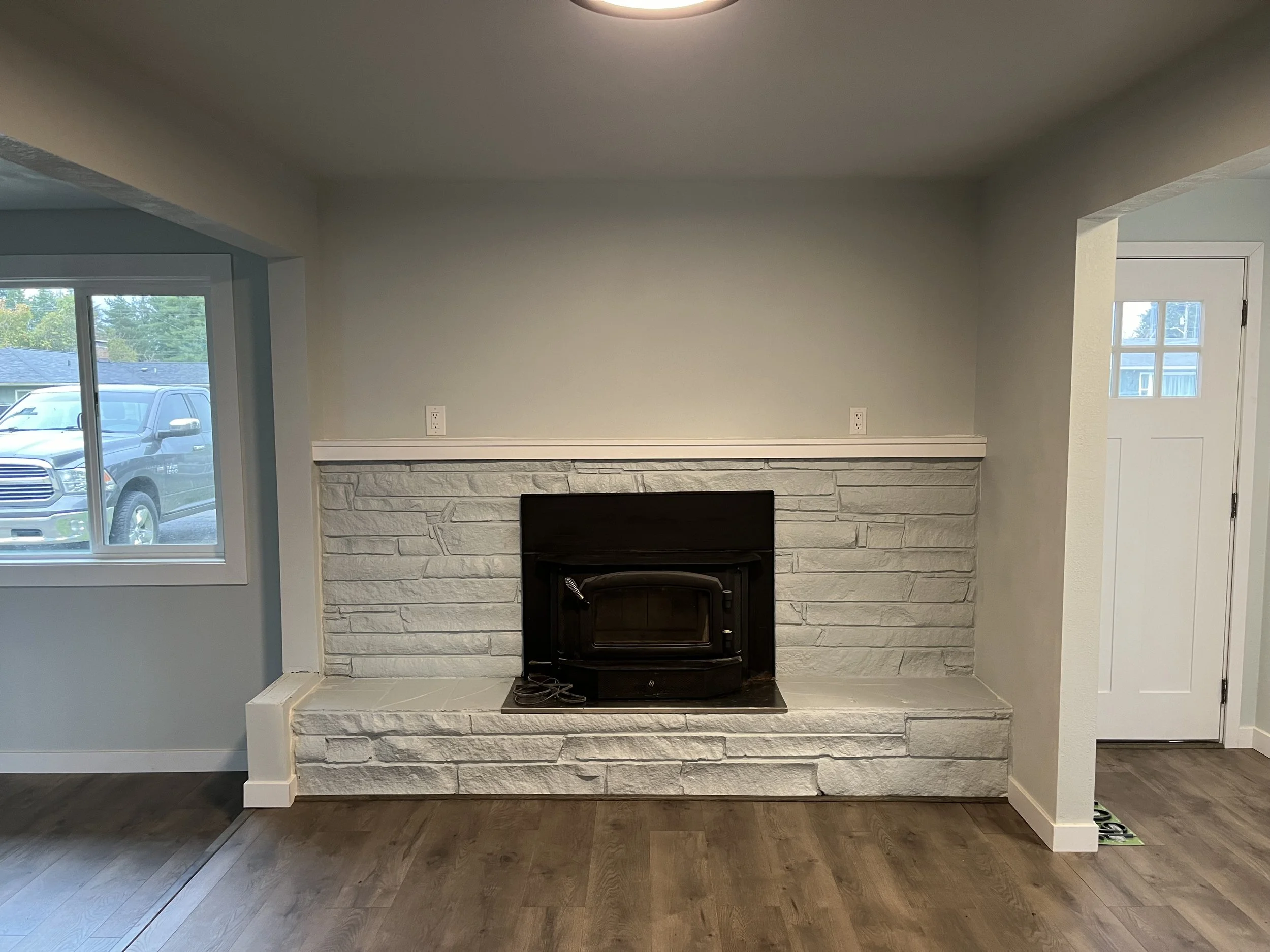 Living room with a white brick fireplace, light gray walls, window with a car outside, and a door on the right side.