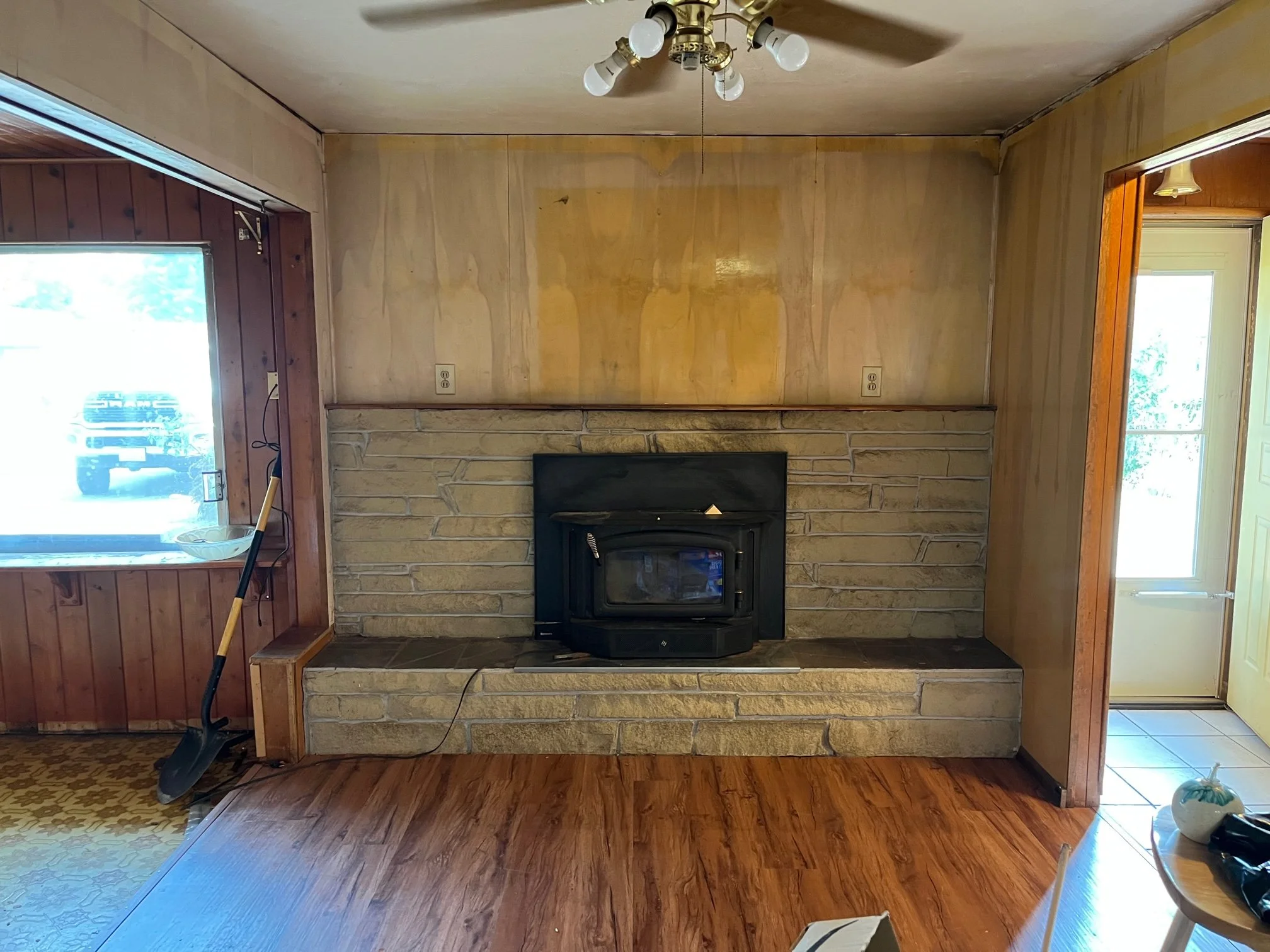 Living room with wood-paneled walls, stone fireplace, and a ceiling fan with light fixtures.