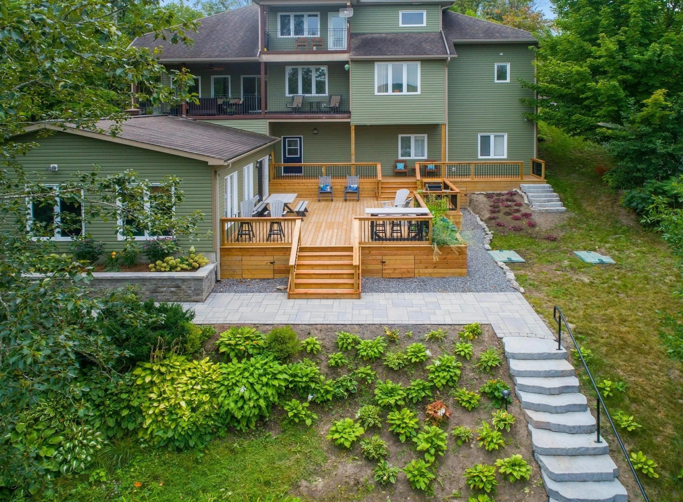 A backyard with a wooden deck, outdoor furniture, and landscaped garden, attached to a multi-story green house surrounded by greenery.