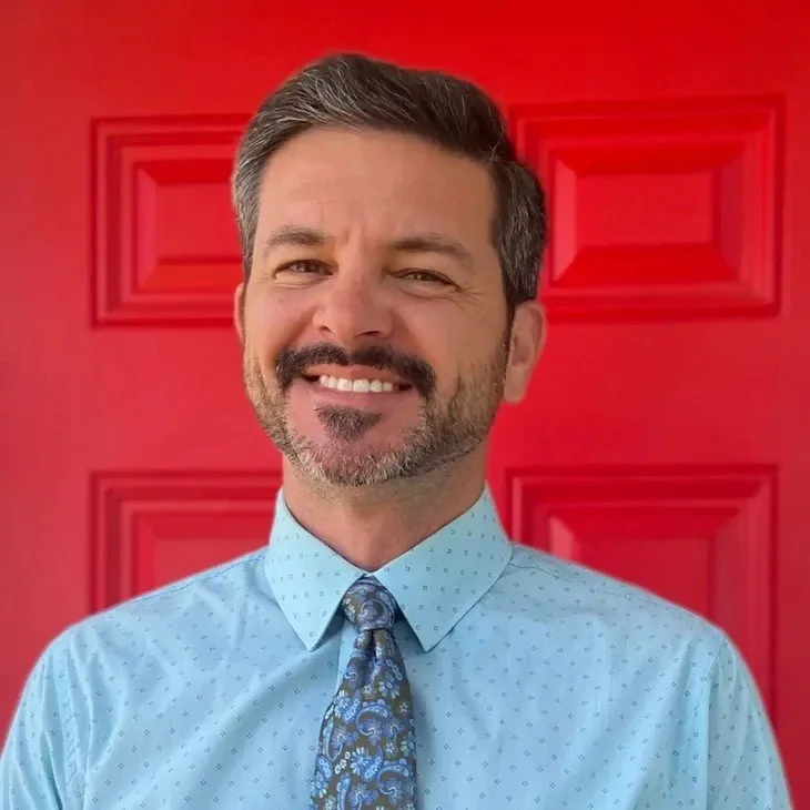 A man with a beard and mustache smiling in front of a red door, wearing a light blue dress shirt and a patterned blue tie.