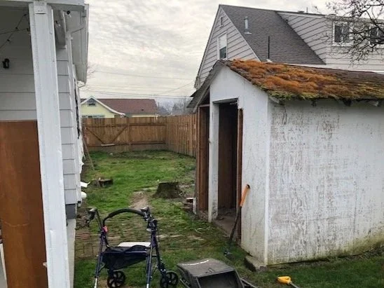 Backyard with a small white shed with moss-covered roof, a fence, and a walker in the yard.