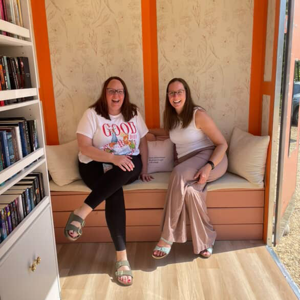 Two women sitting on a peach-colored cushioned bench in a cozy, well-lit room with bookshelves to the side and orange striped decor on the wall, smiling and posing for the photo.