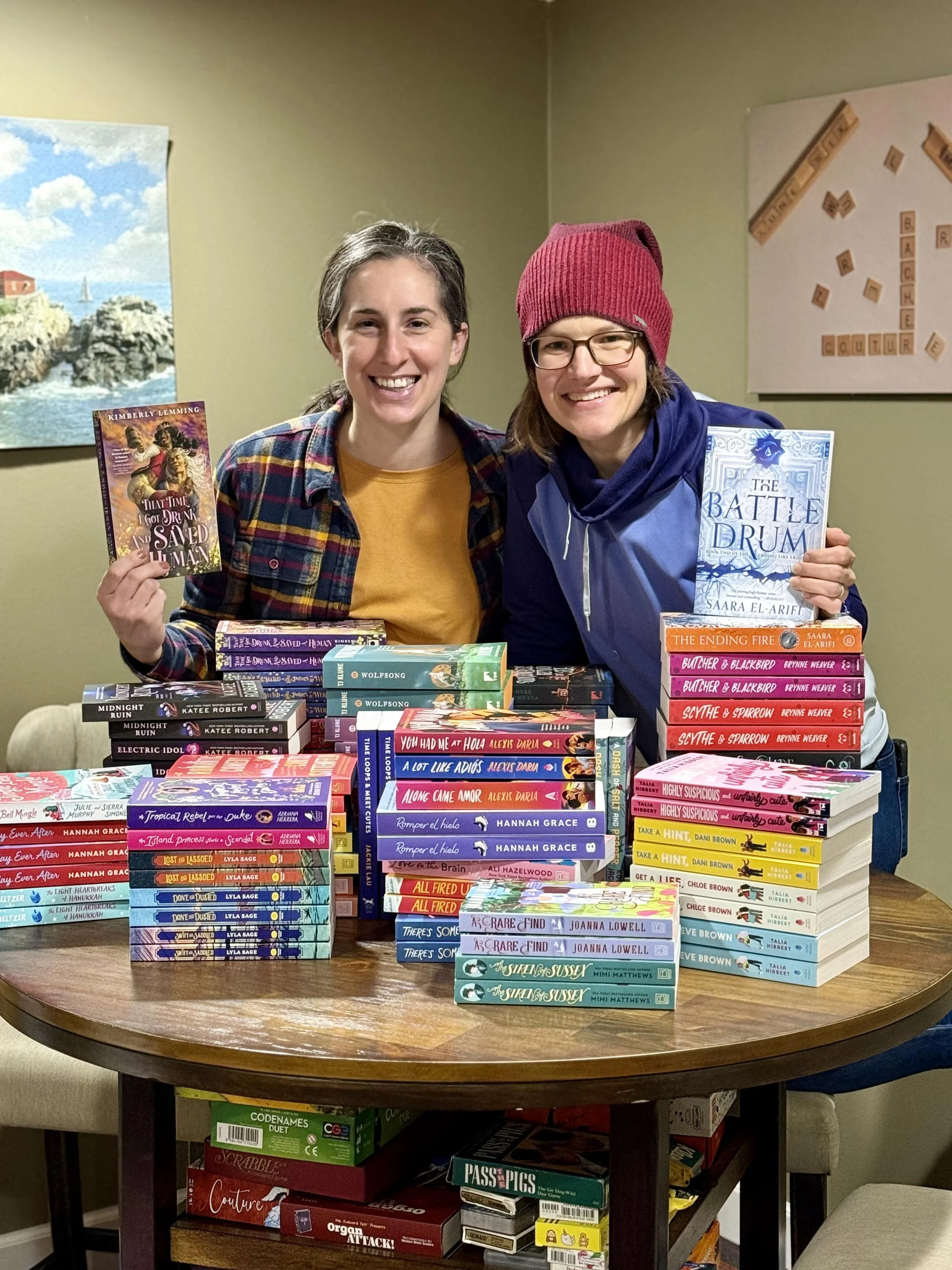 Two smiling women standing behind a table filled with stacks of colorful books, holding one book each, inside a room with green walls, paintings, and a board with wooden letter tiles.