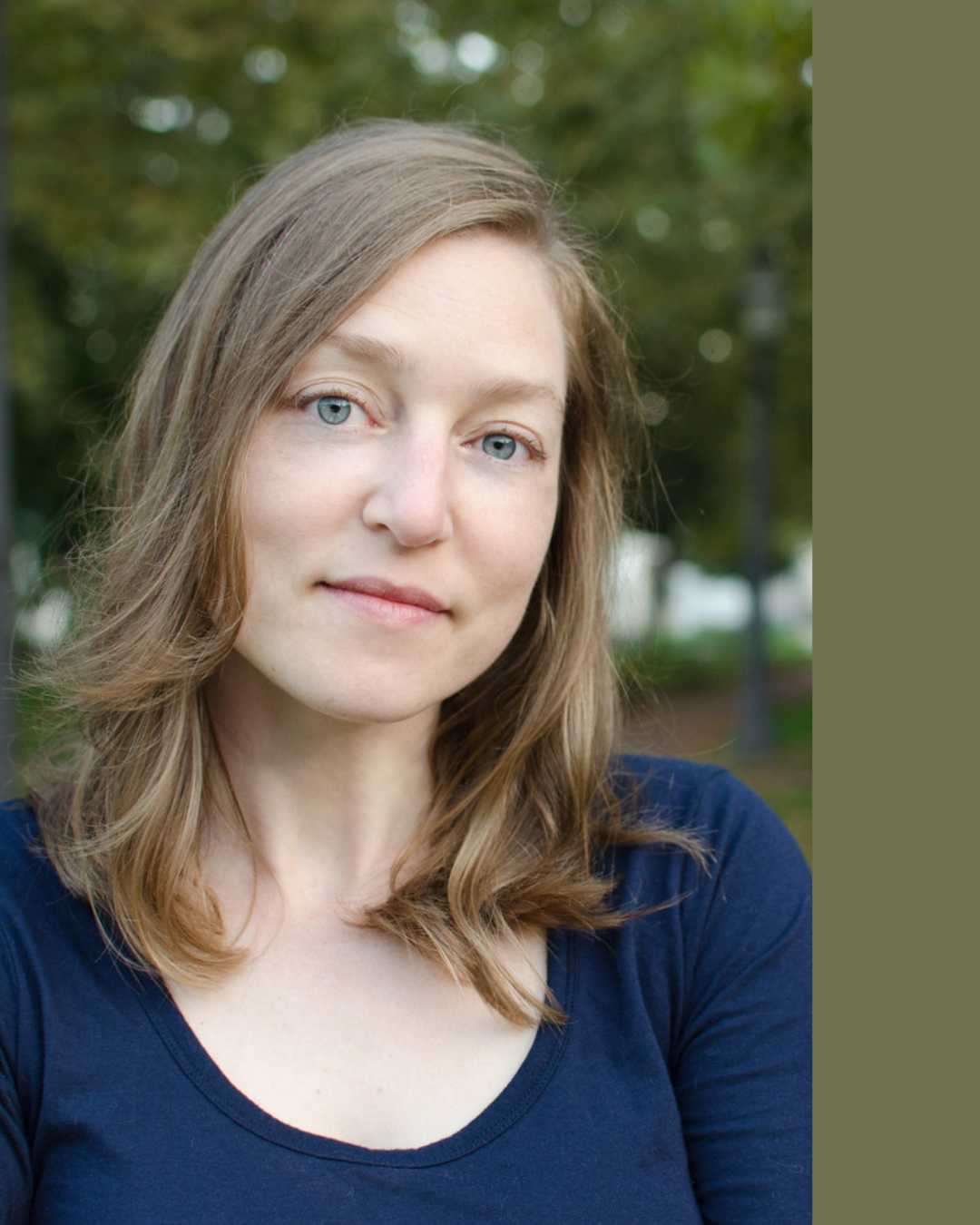 Close-up of a young woman with light brown hair and blue eyes, wearing a navy blue shirt, standing outdoors with trees in the background.
