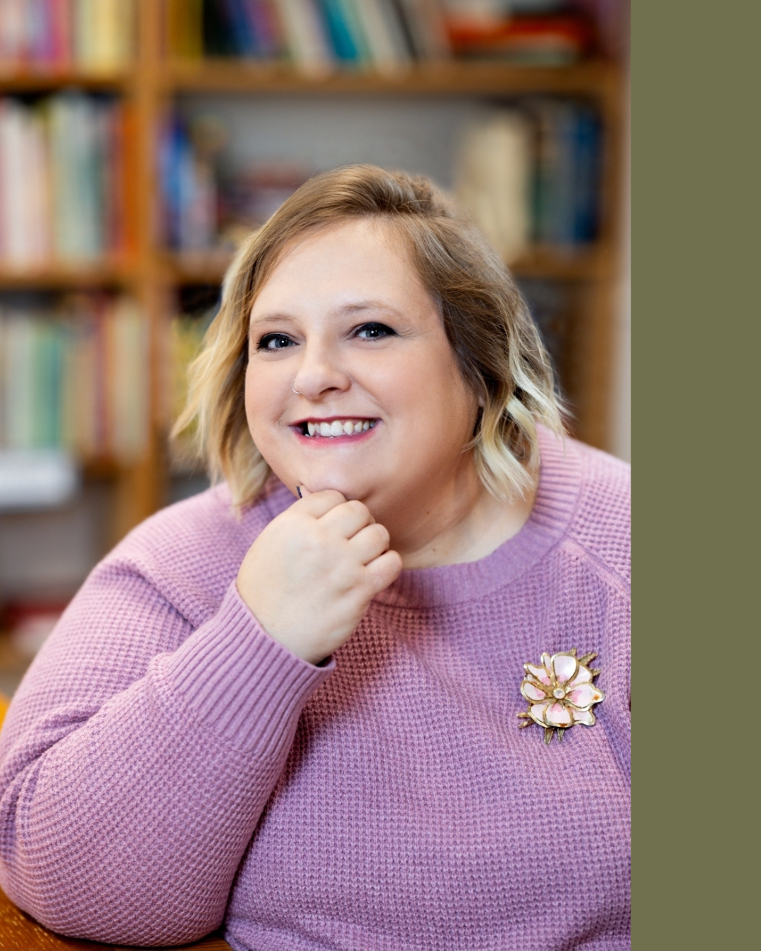 A smiling woman with short blonde hair, wearing a pink sweater and a floral pin, sitting in front of a bookshelf.