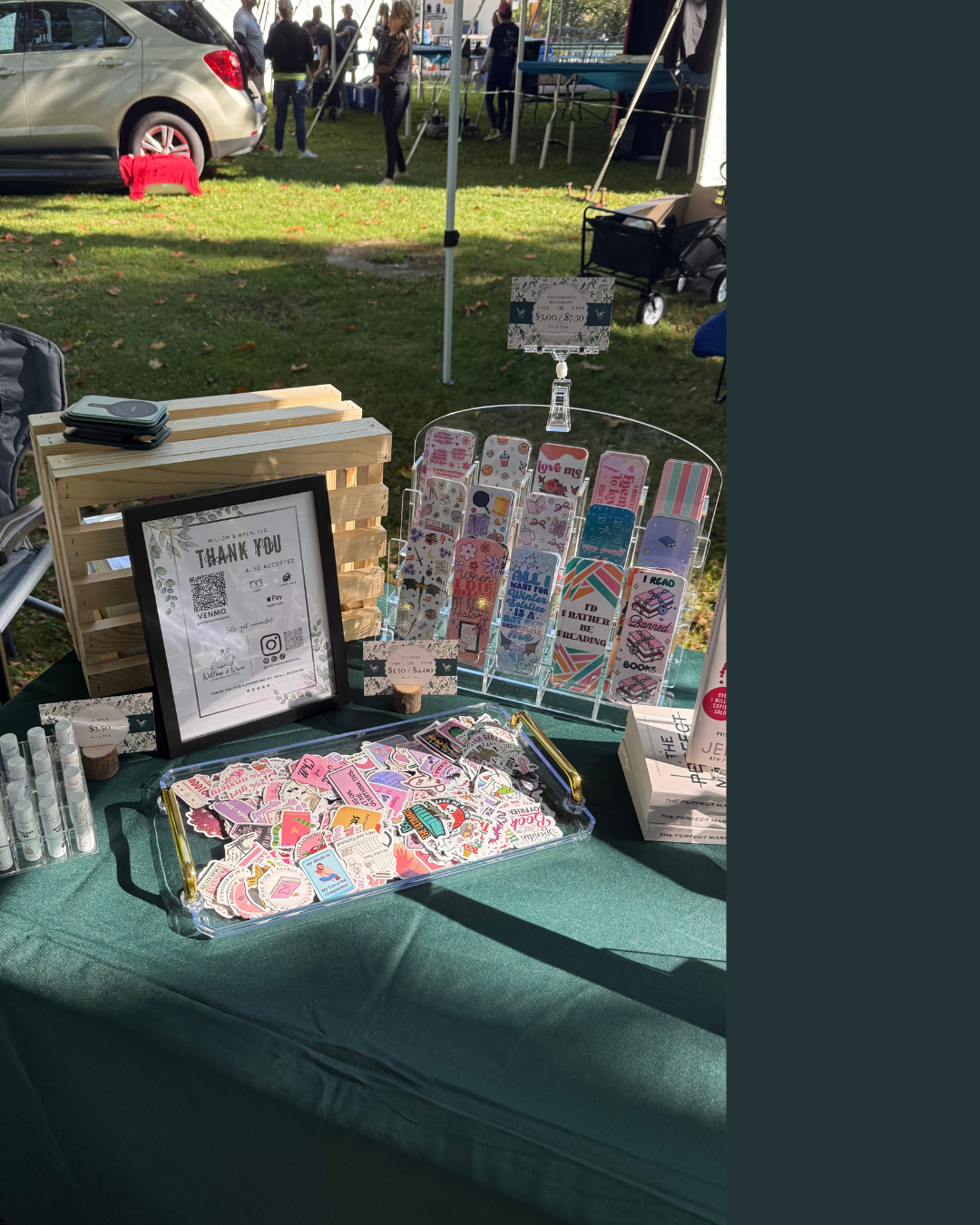 A display table at an outdoor market selling colorful stickers, greeting cards, and small paper goods. The table has a framed thank you sign, a tray filled with various stickers, and stands showcasing bookmarks and small cards. There are small cylindrical containers on the left, and in the background, people are walking and standing under tents.