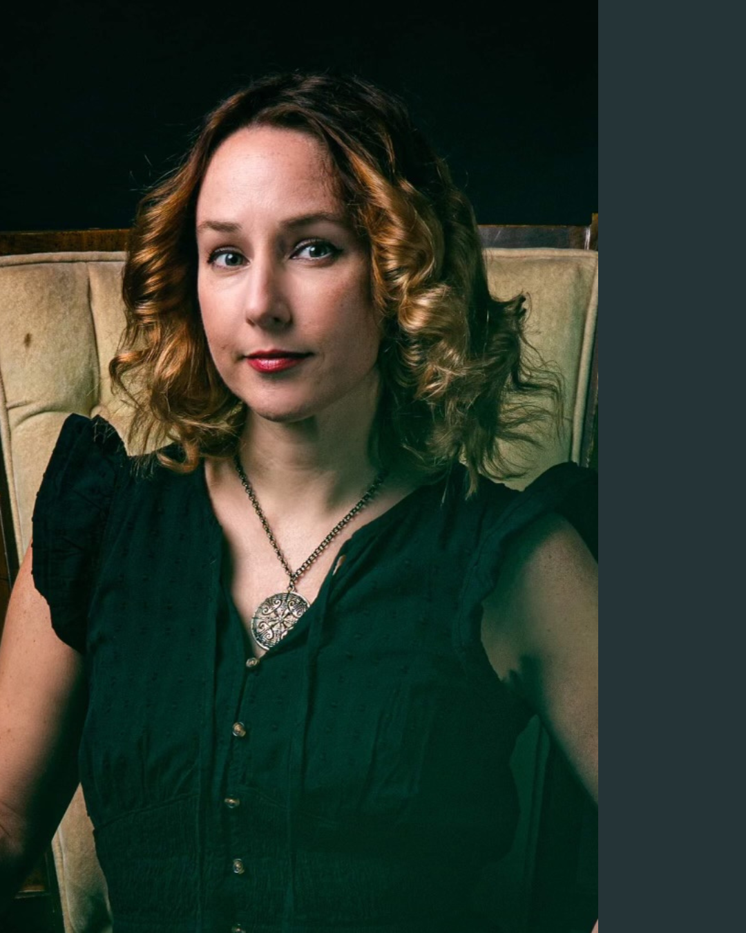 A woman with curly, shoulder-length reddish hair, wearing a black top and a silver pendant necklace, sitting against a beige and brown cushioned background.