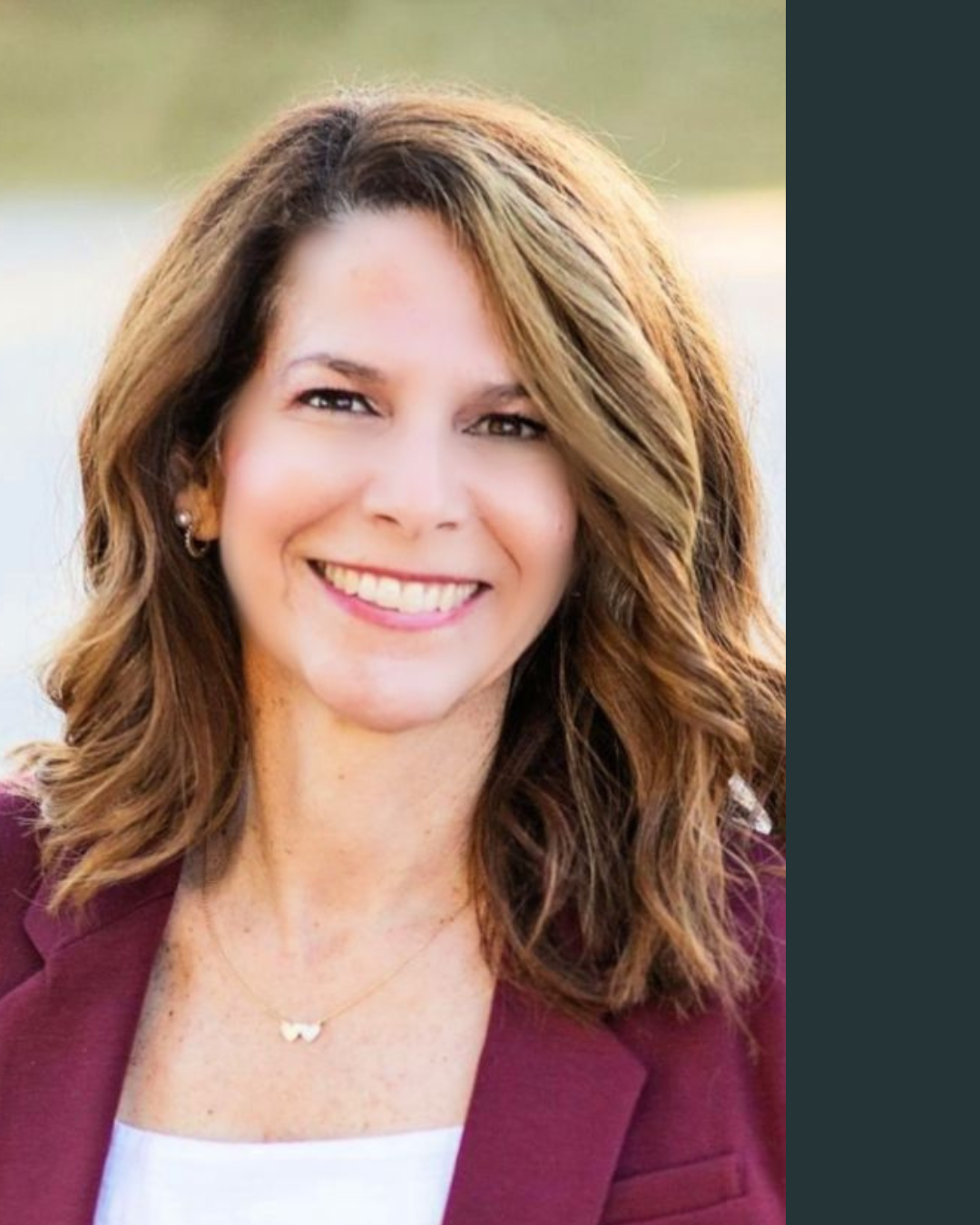 Close-up of a smiling woman with shoulder-length wavy brown hair, wearing a burgundy blazer and a white top, outdoors with blurred background.