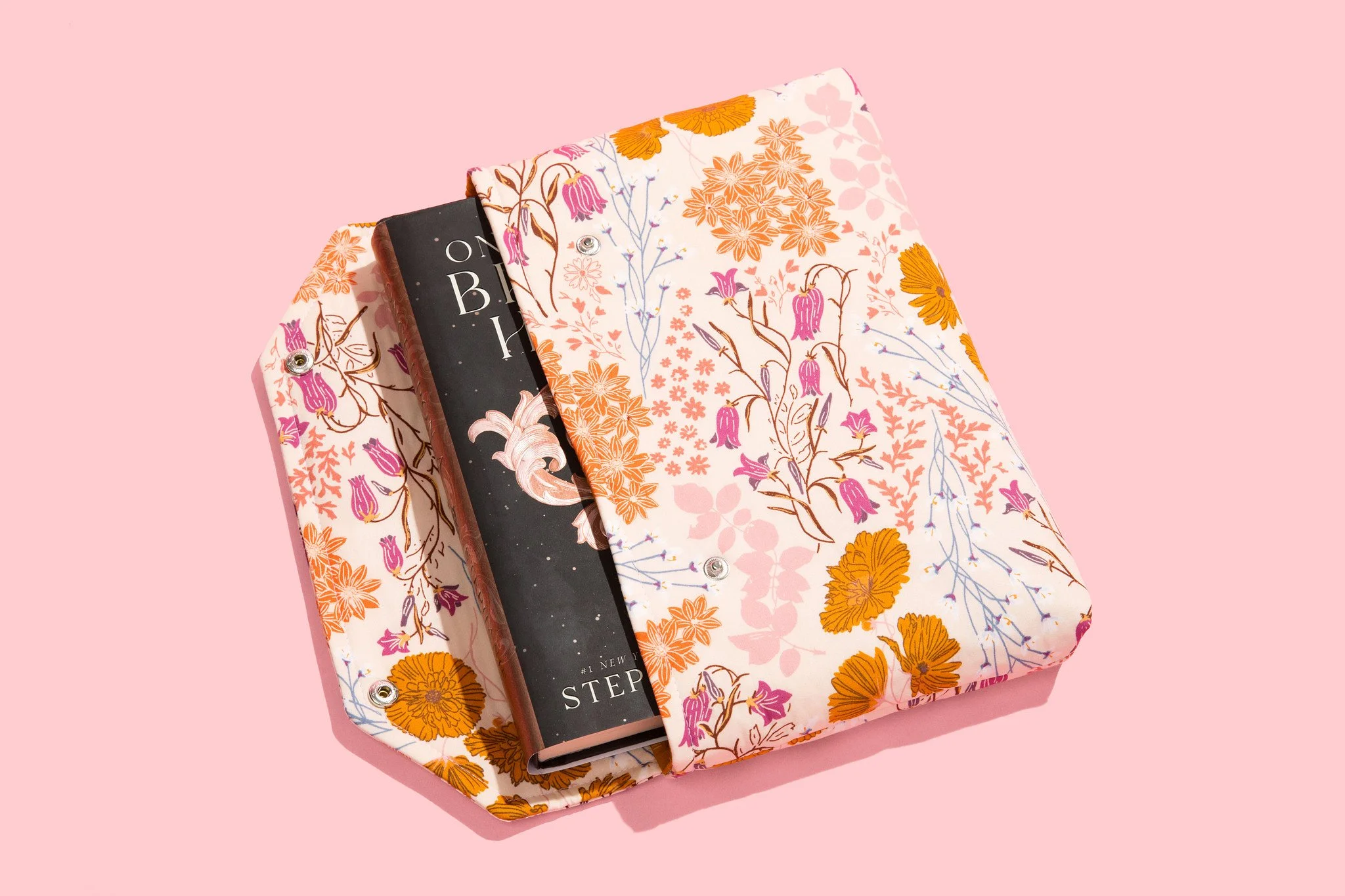 A floral-patterned fabric bag with a book partially inside, set against a pink background.