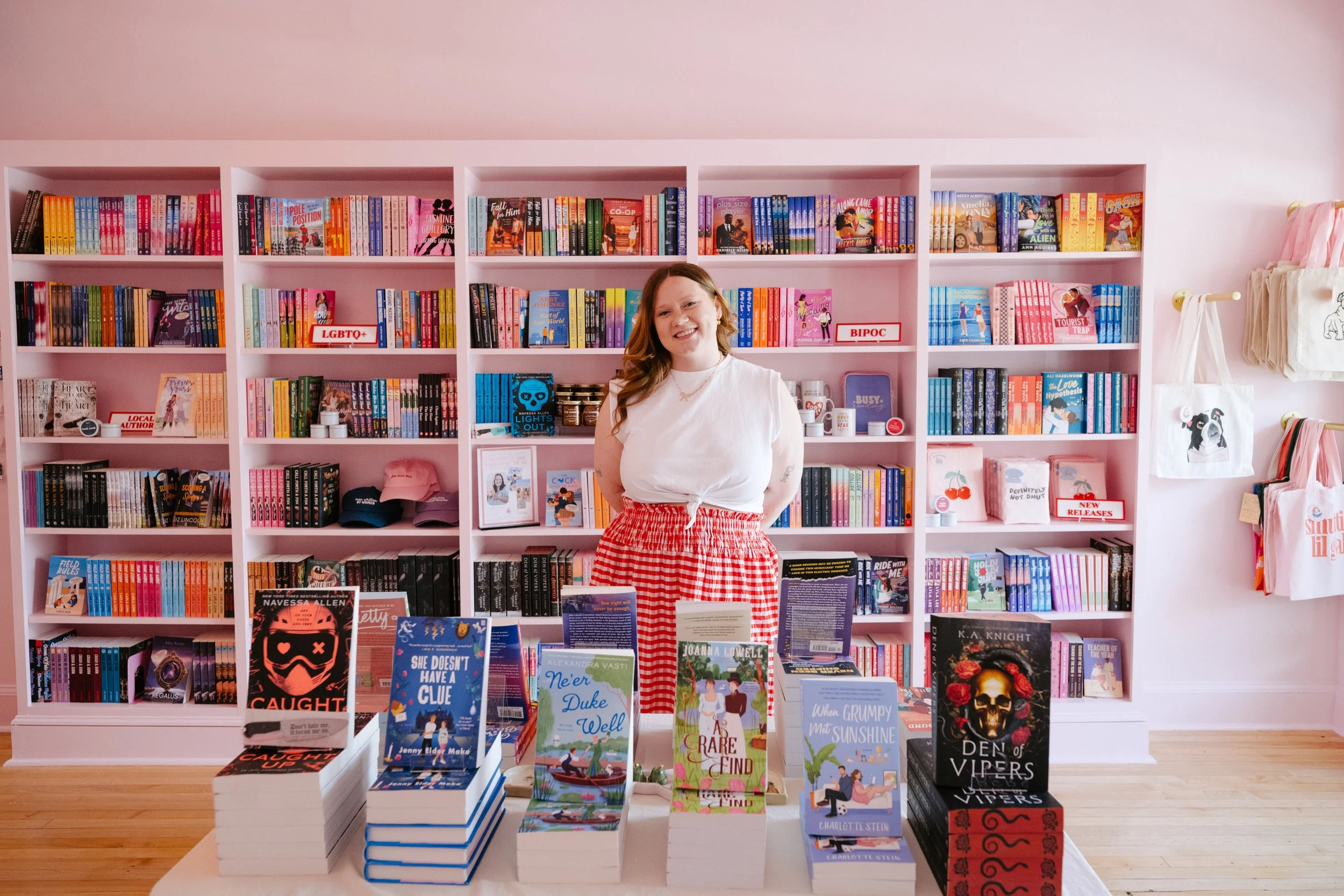 Woman standing in front of bookshelves in a bookstore, smiling, wearing a white top and red checkered skirt, surrounded by stacks of books on a display table.