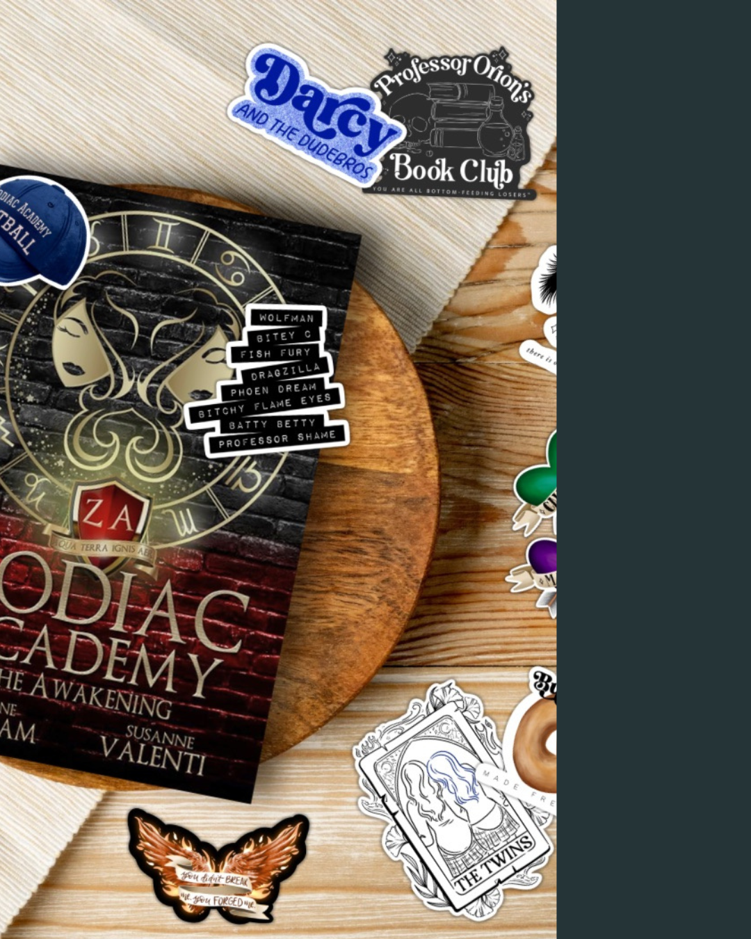 A wooden table with various stickers and a copy of the book "Zodiac Academy: The Awakening" by Susanne Valenti partially visible, surrounded by stickers including a black and white one for "Professor Orion's Book Club", a blue sticker for "Dacy and the Dudebros", a sticker with a drawing of twins, and various other decorative stickers.