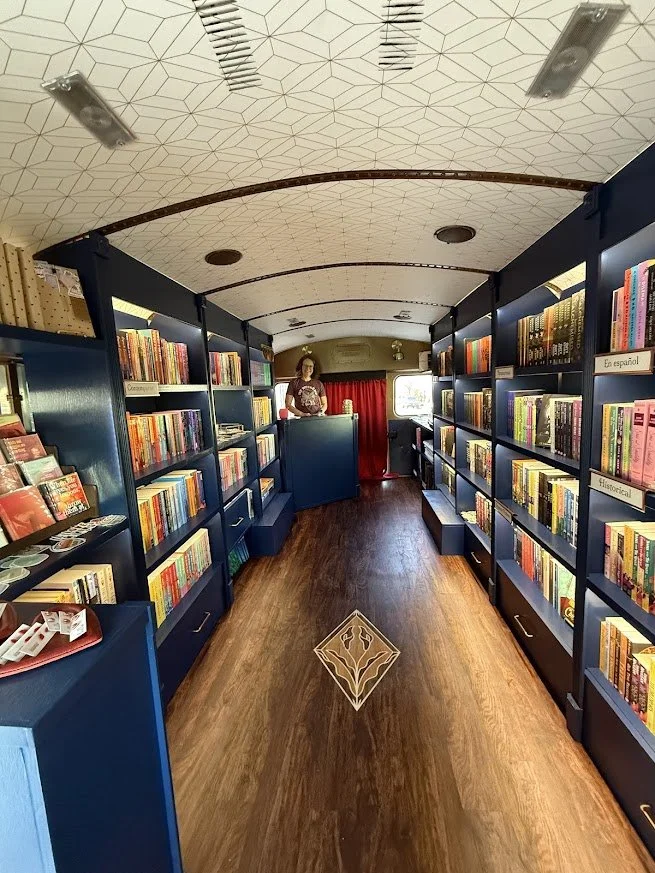 A small bookstore with dark blue bookshelves filled with colorful books. A woman stands behind a small counter near a red curtain, with a window and natural light in the background. The ceiling is decorated with a geometric pattern and has lighting fixtures. The wooden floor has a decorative inlay in the center.