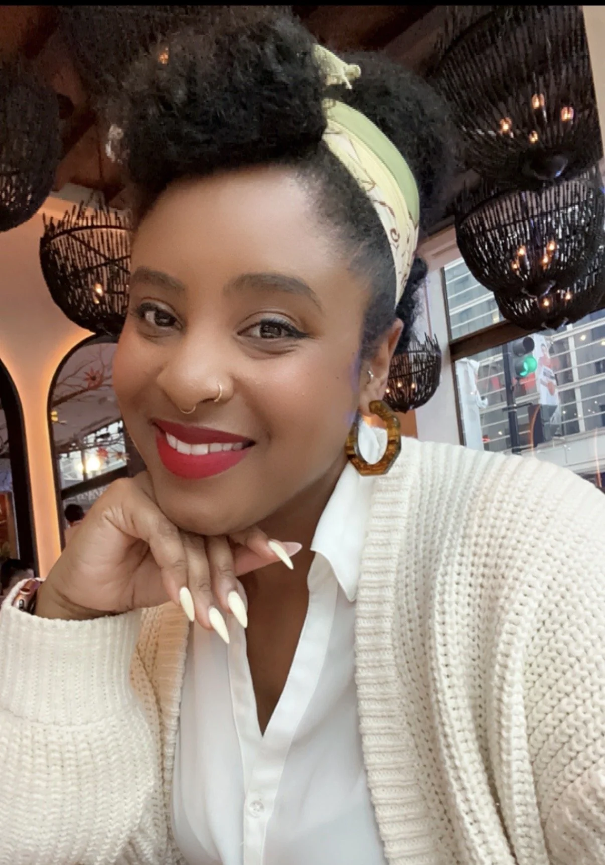 A smiling woman with short curly hair in a cream sweater, wearing red lipstick, hoop earrings, and a headband, inside a restaurant or cafe with black woven pendant lights and large windows.