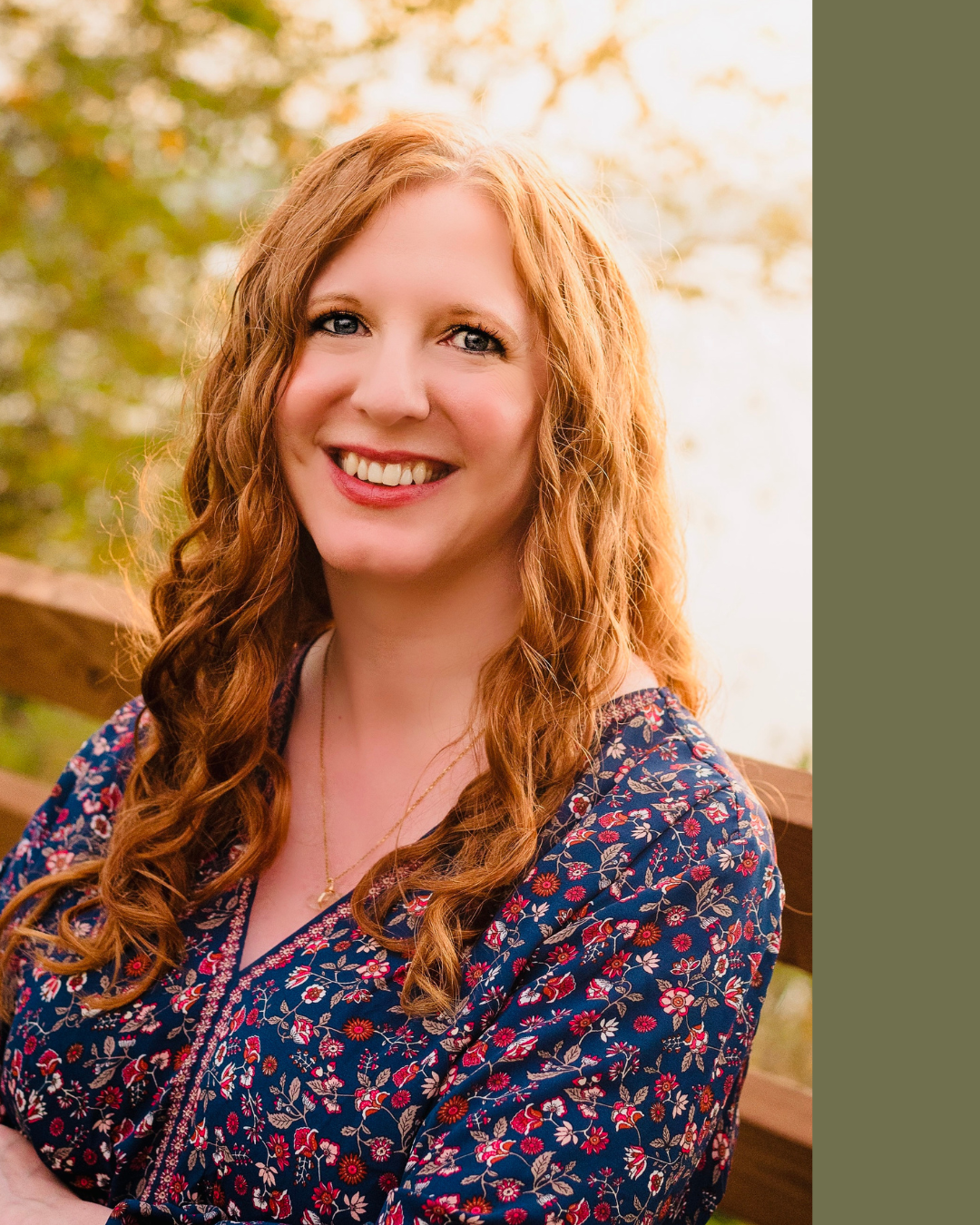 A smiling woman with curly red hair, wearing a floral dress, outdoors with trees and a wooden fence in the background.