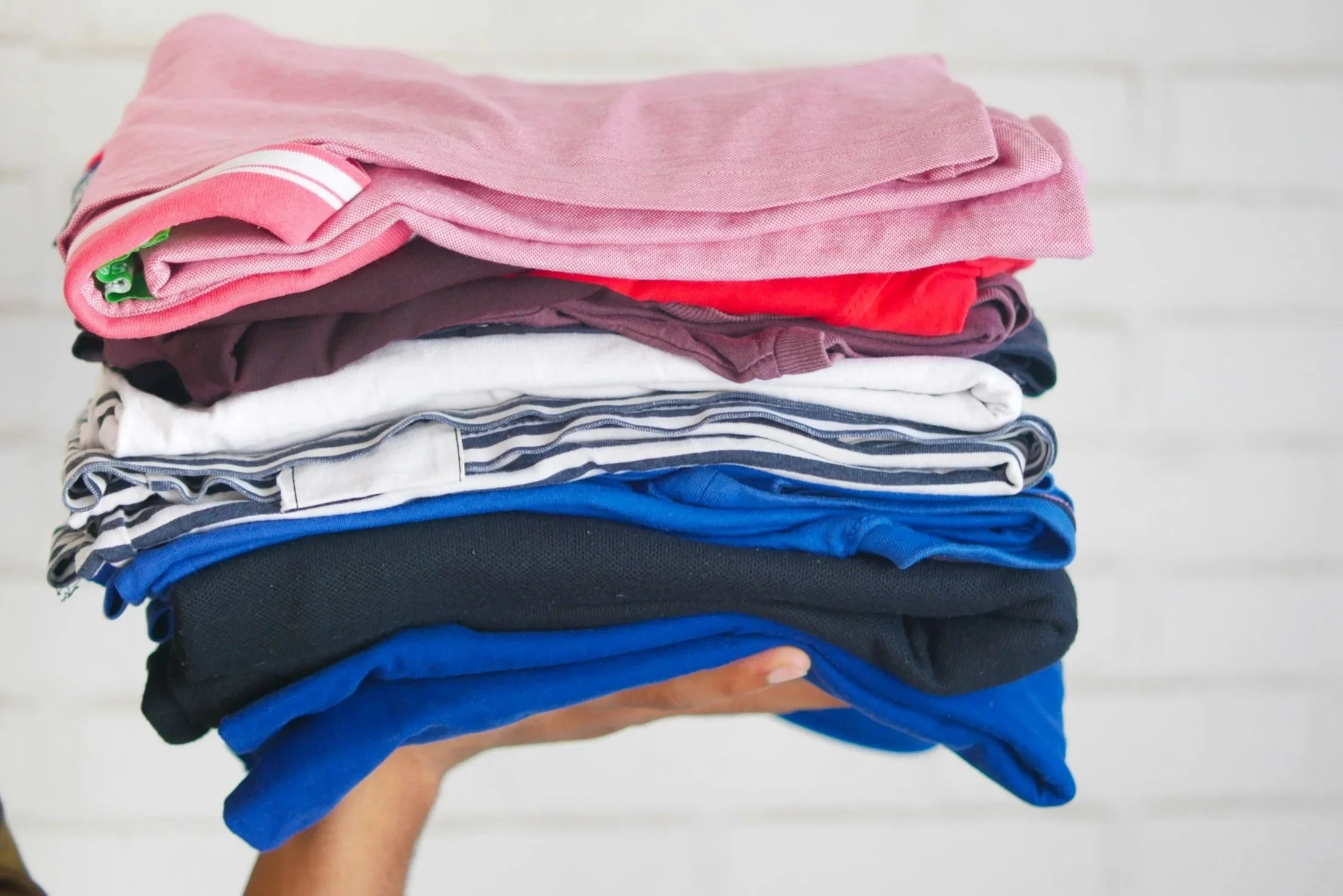 A hand holds a stack of folded clothes, including pink, red, white with blue stripes, black, and blue shirts, against a white background.