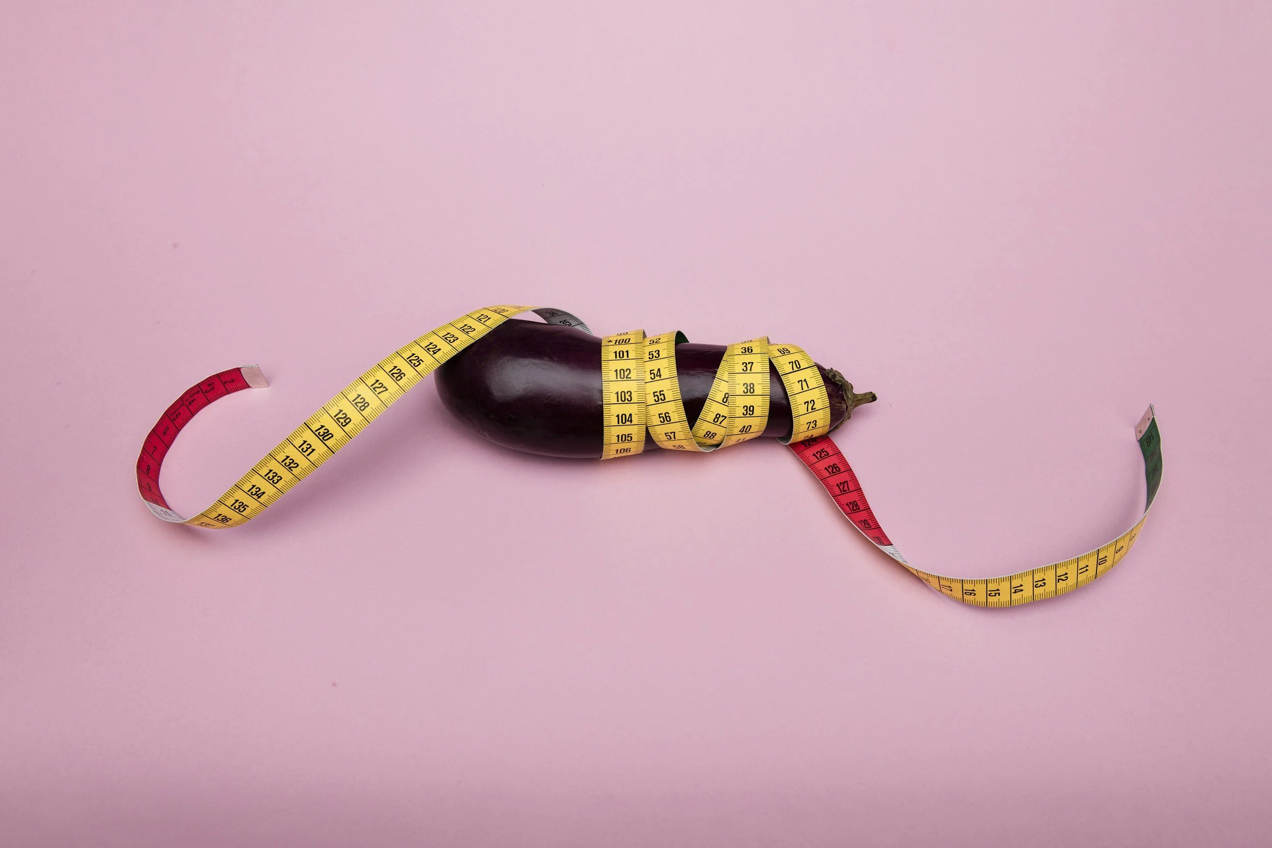 An eggplant wrapped with a yellow measuring tape, lying on a pink background.