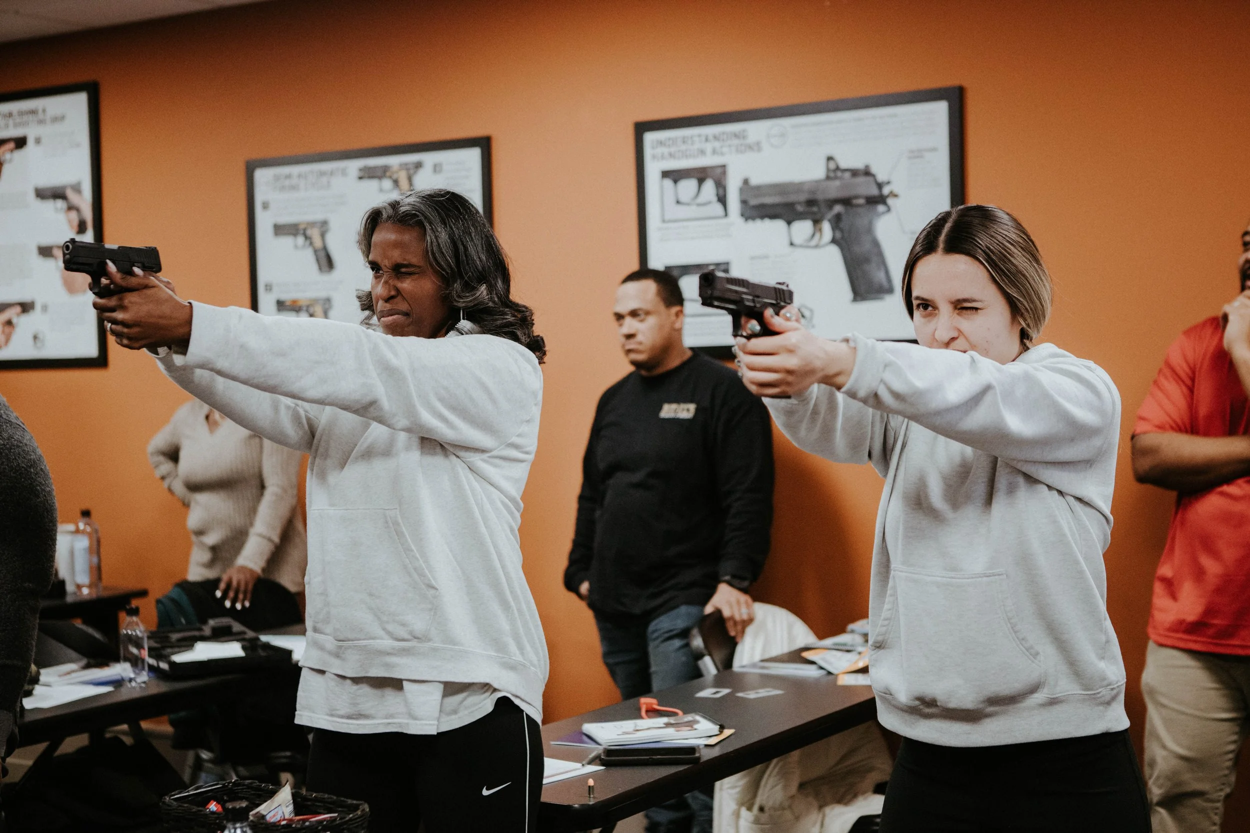 Two women aiming guns during a training class with posters of firearms on the orange wall behind them.