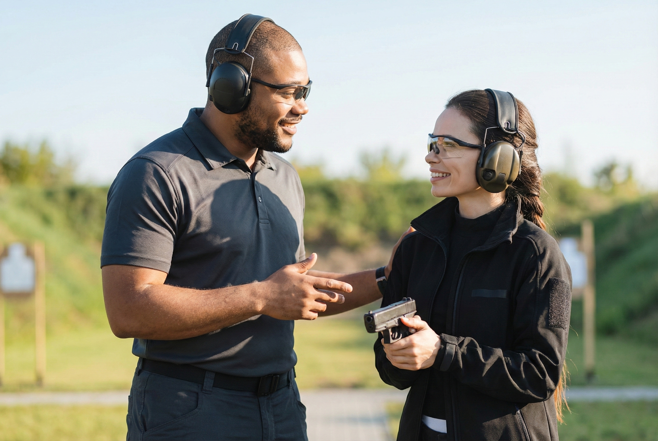 Man and woman at outdoor shooting range, wearing hearing protection and safety glasses, woman holding a handgun, man talking to her.