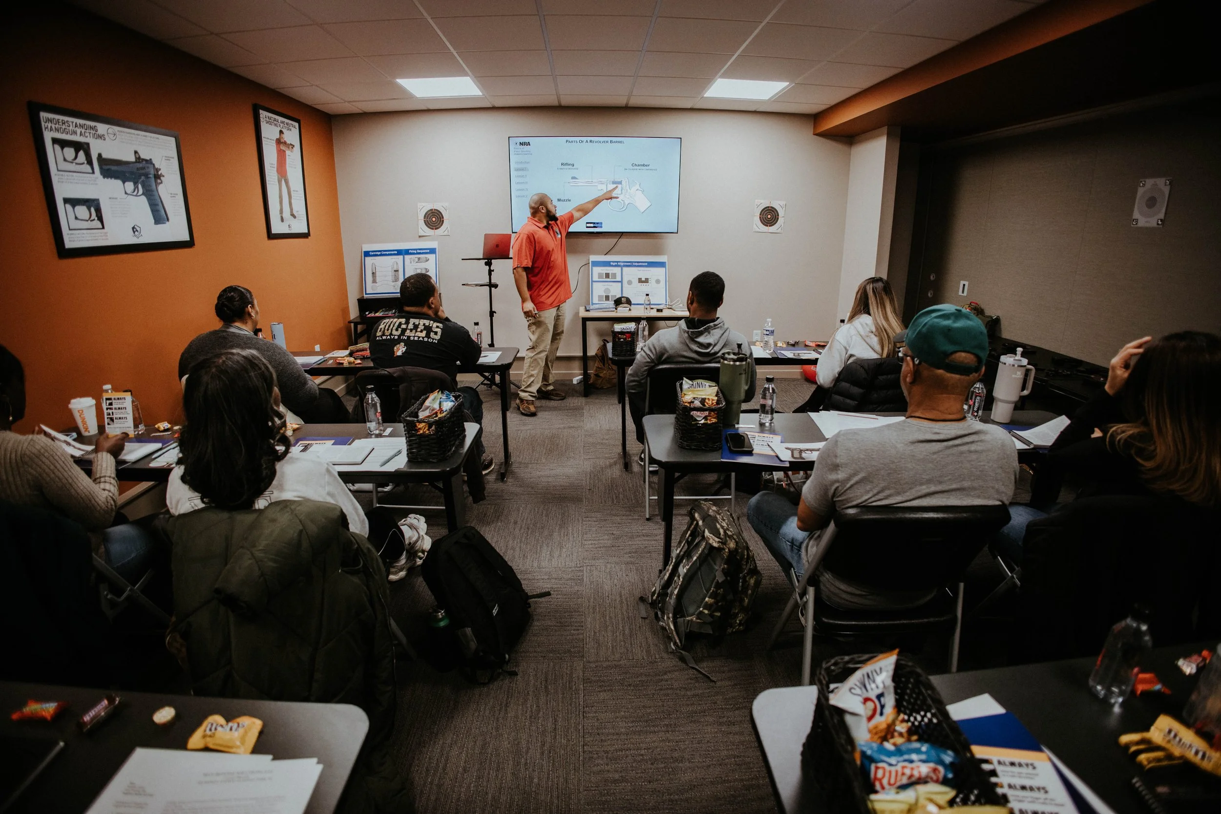 A classroom with a teacher at the front pointing at a screen, students seated at desks, some with backpacks and snacks, in a training session on firearms.