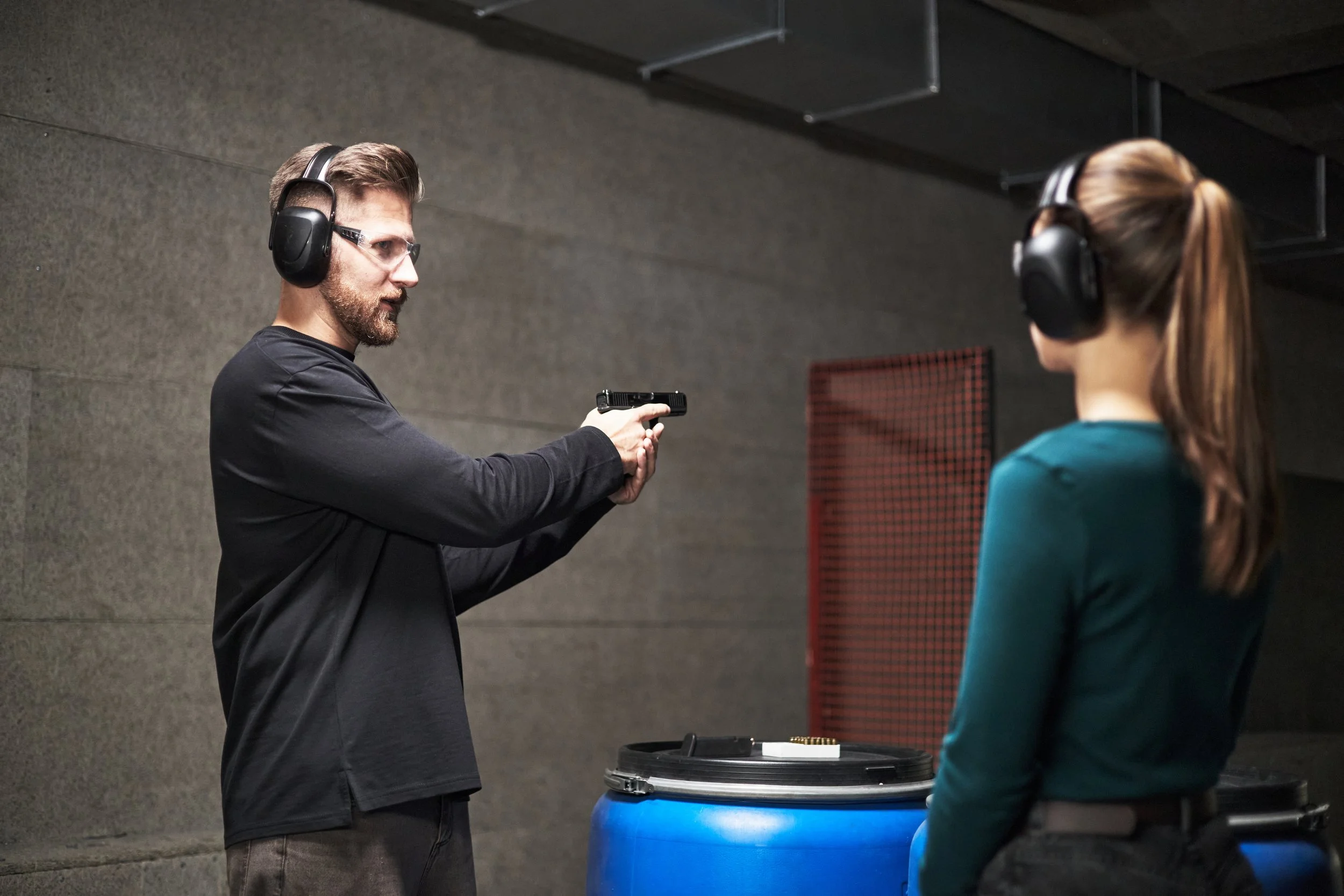 A man in black clothing pointing a handgun at a woman in a shooting range, both wearing hearing protection.