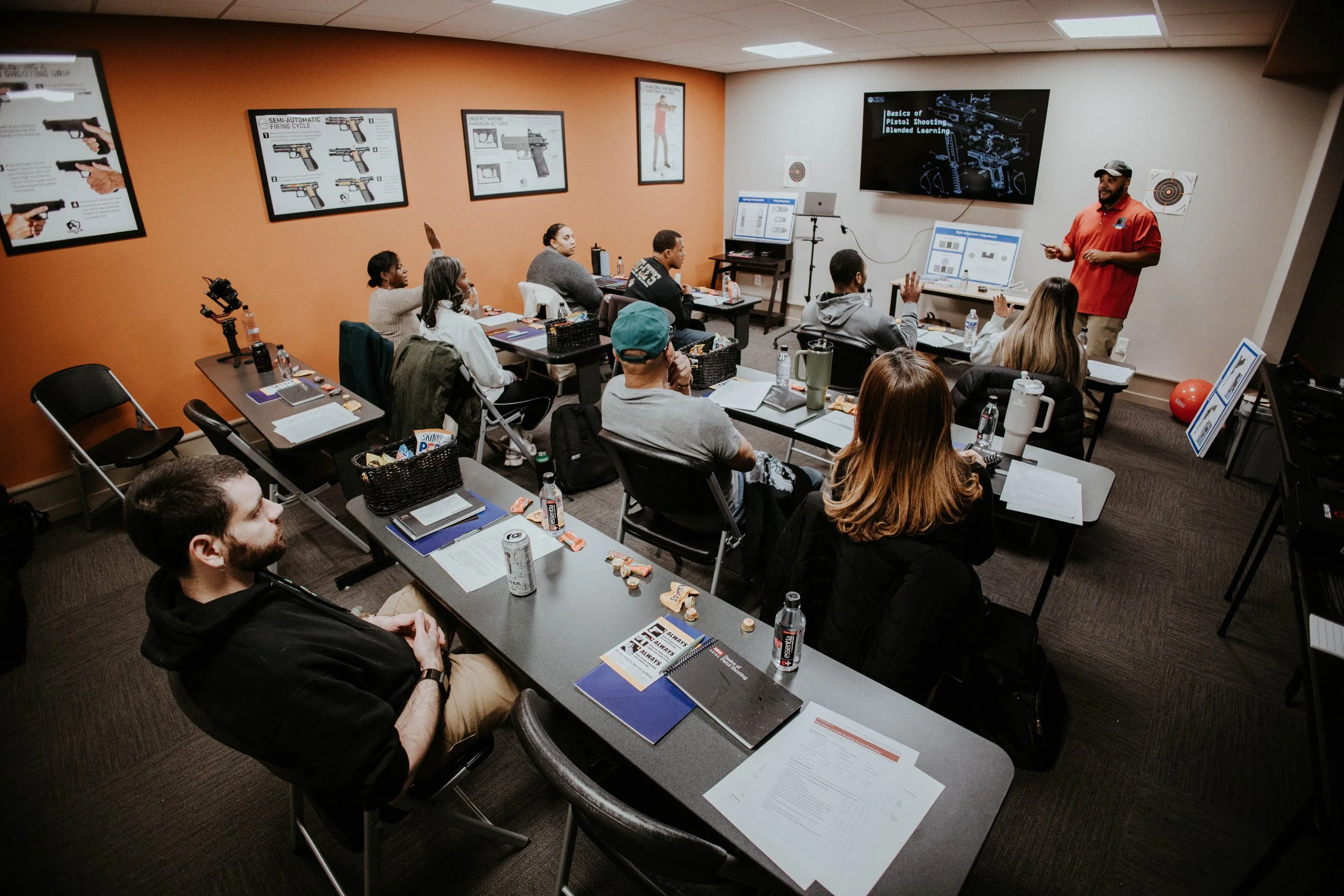 A classroom with students attending a firearm safety or gun handling training session, with instructor at the front, presentation materials, and posters on firearm diagrams on the walls.