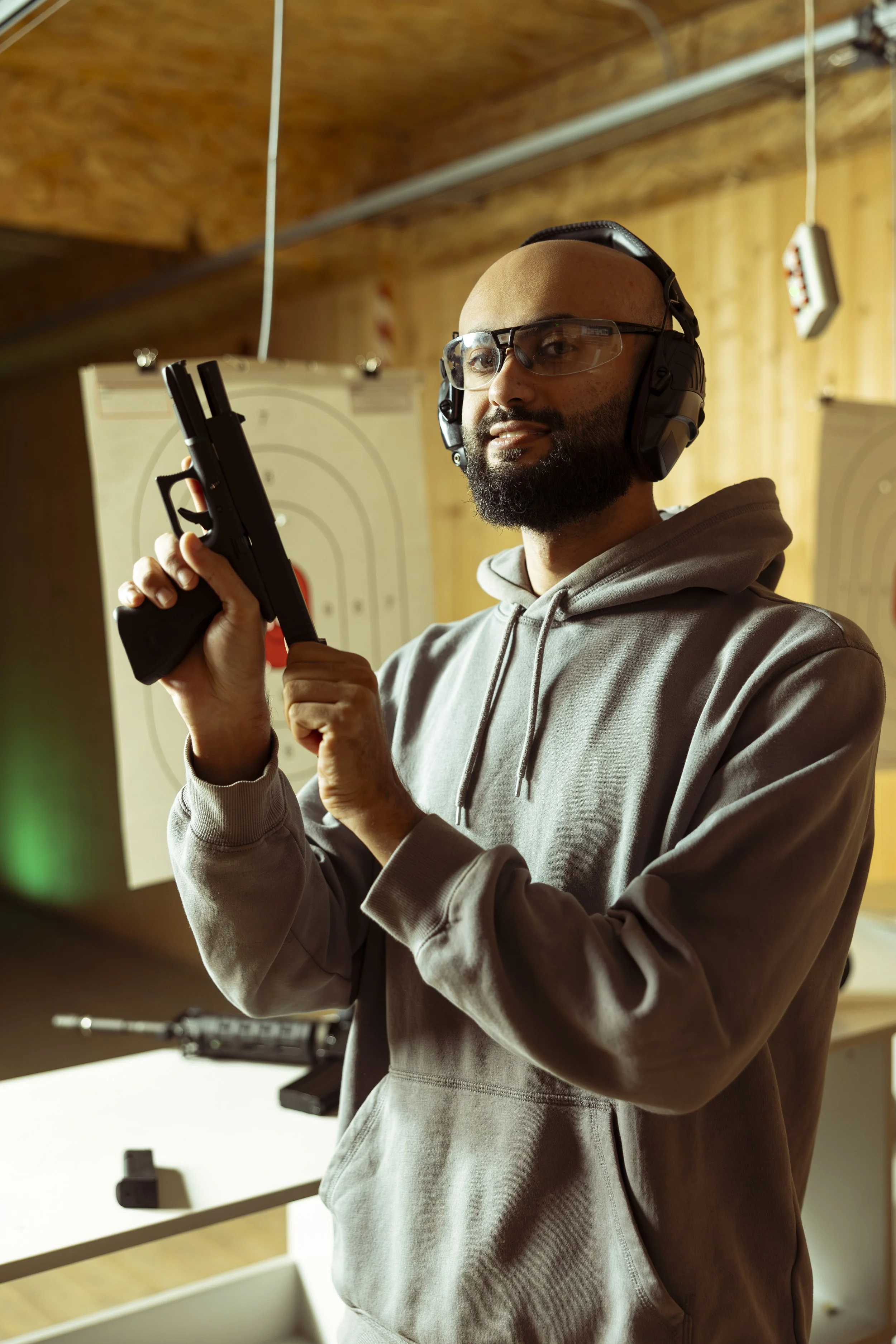 A man in safety glasses and headphones holding a handgun at an indoor shooting range with target boards in the background.