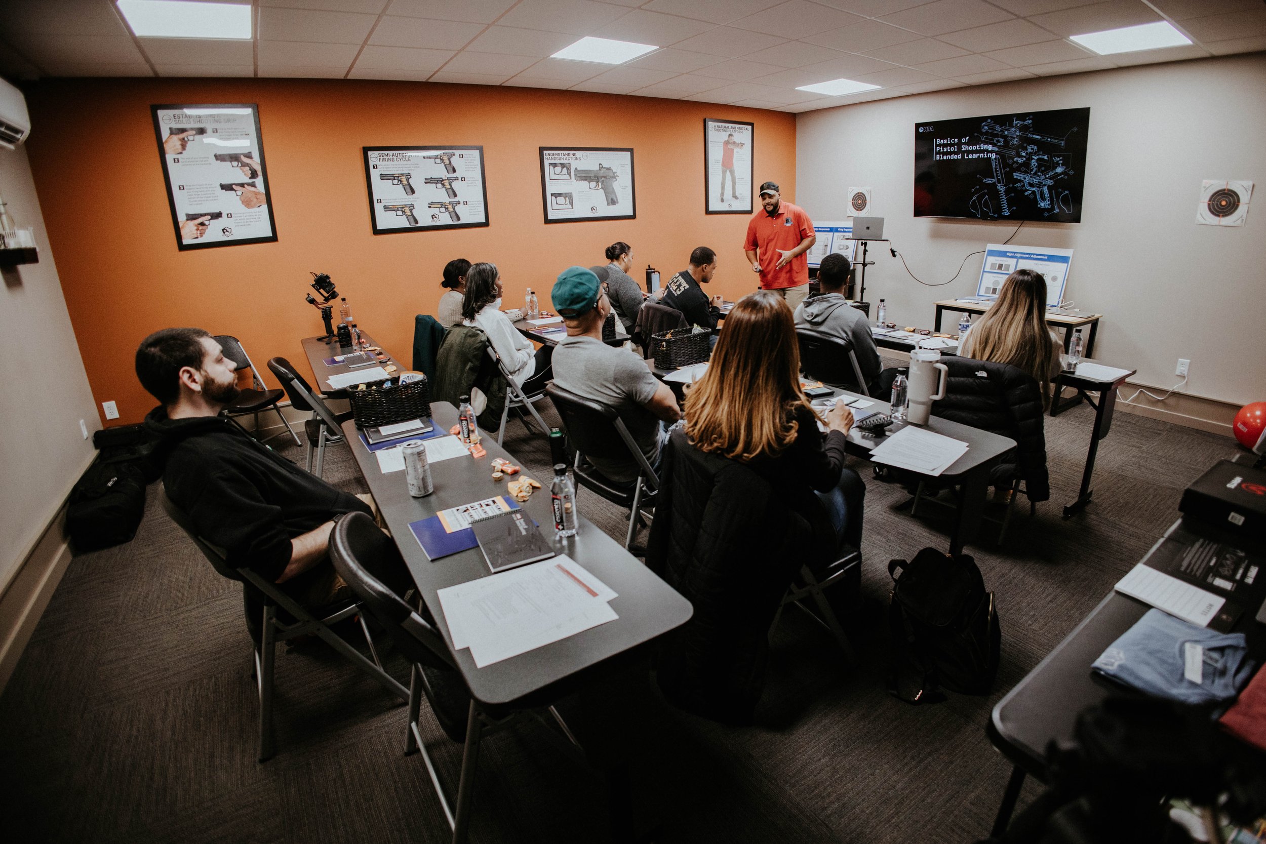 Indoor classroom or workshop setting with attendees seated at tables, listening to a speaker at the front who is demonstrating firearm techniques using a large screen and posters on the wall related to guns and shooting.