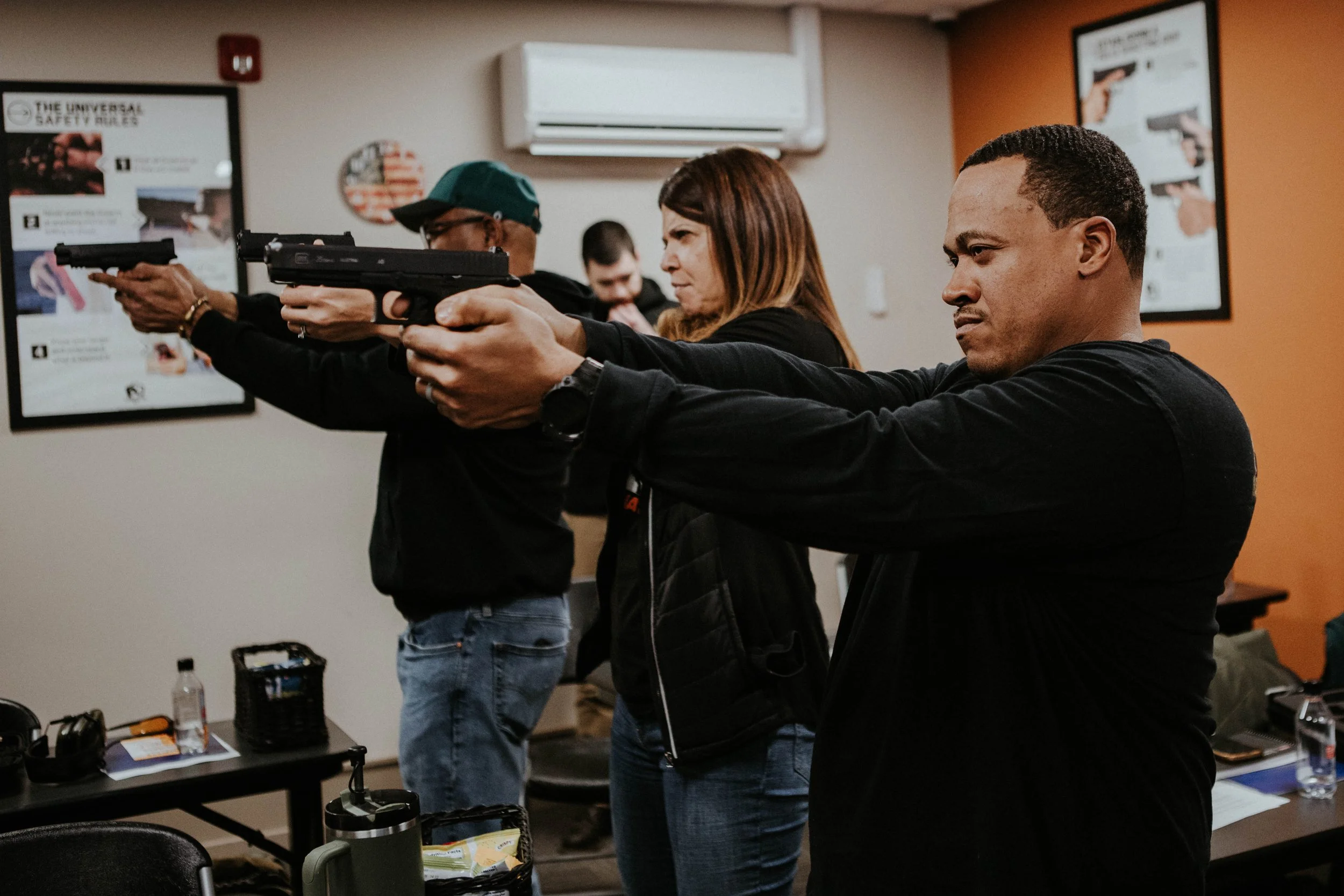 Three people practicing firearm safety and handling, aiming handguns in a classroom with safety posters on the wall.