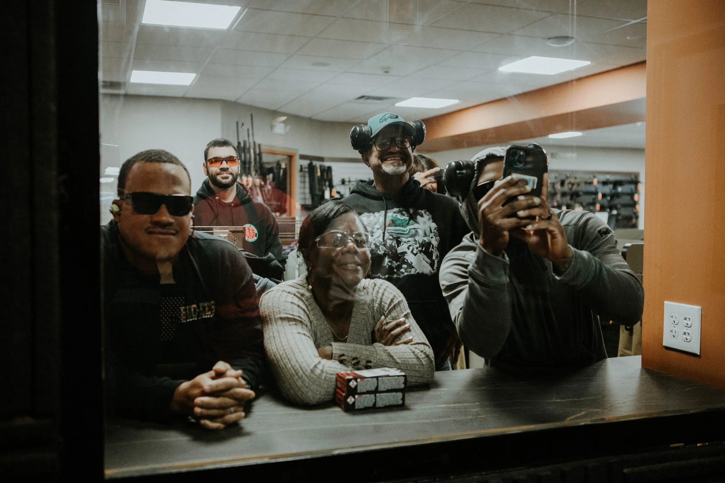 A group of five people behind a window, taking a photo or video of themselves in a store or workshop with firearms and ammunition on display in the background.