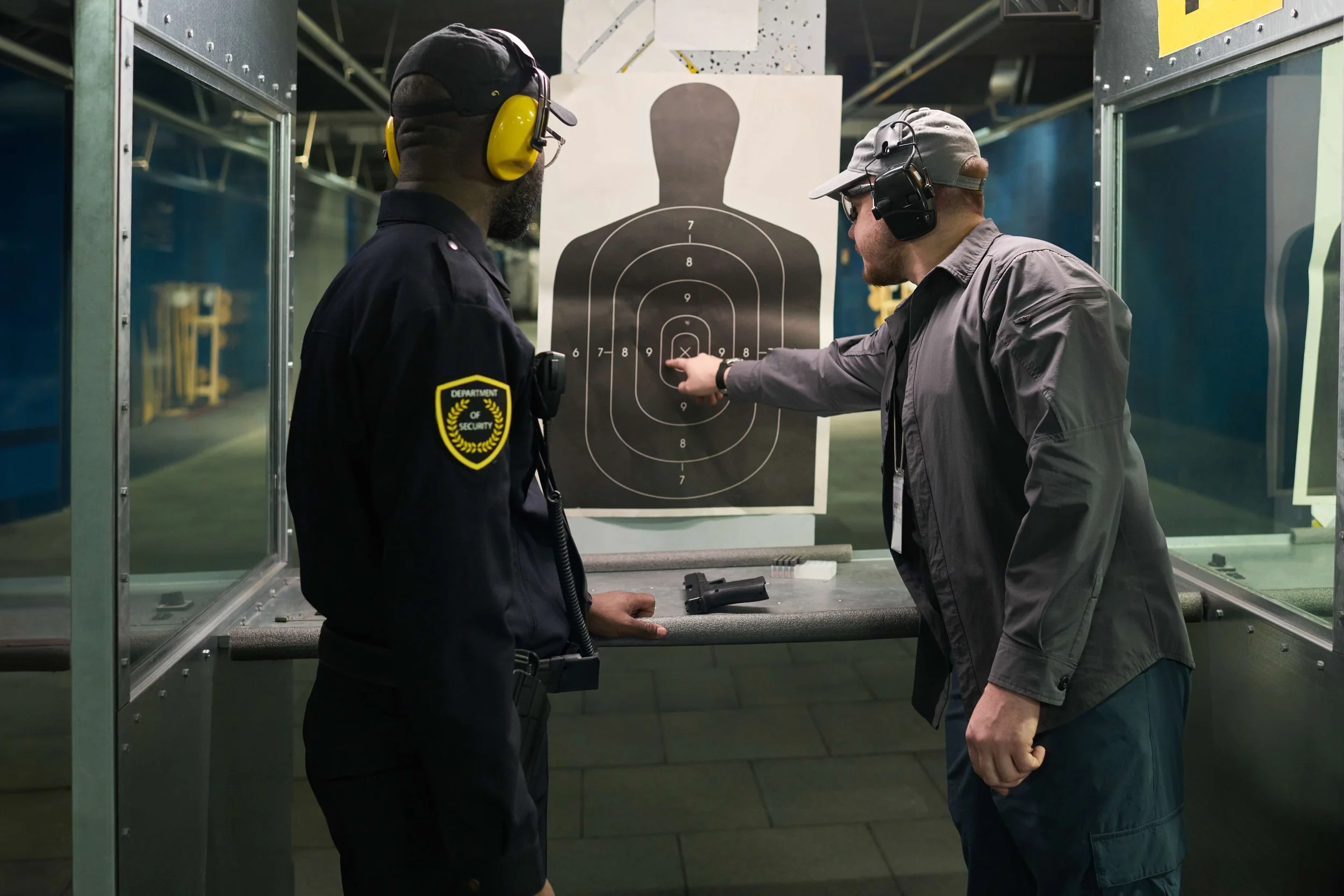 A security officer and a man at a shooting range, with a target featuring a human silhouette on a stand between them. The man is pointing at the target's chest area while the officer looks on.