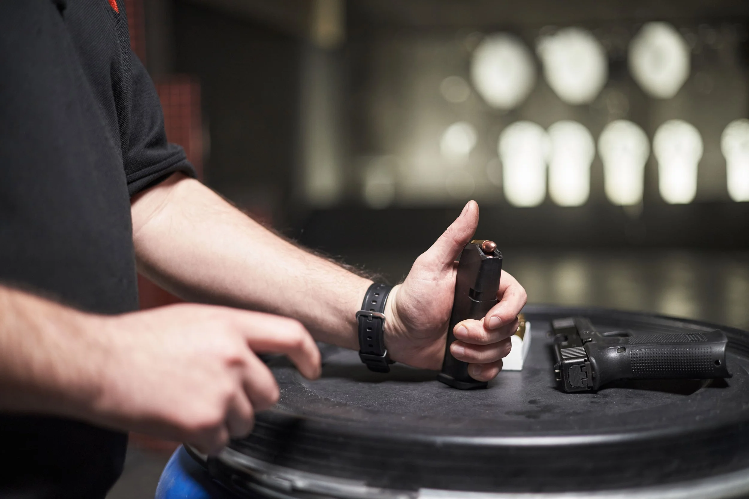 A person holding a handgun with a magazine inserted, next to another handgun on a black surface, with blurred background showing a wall with multiple windows.