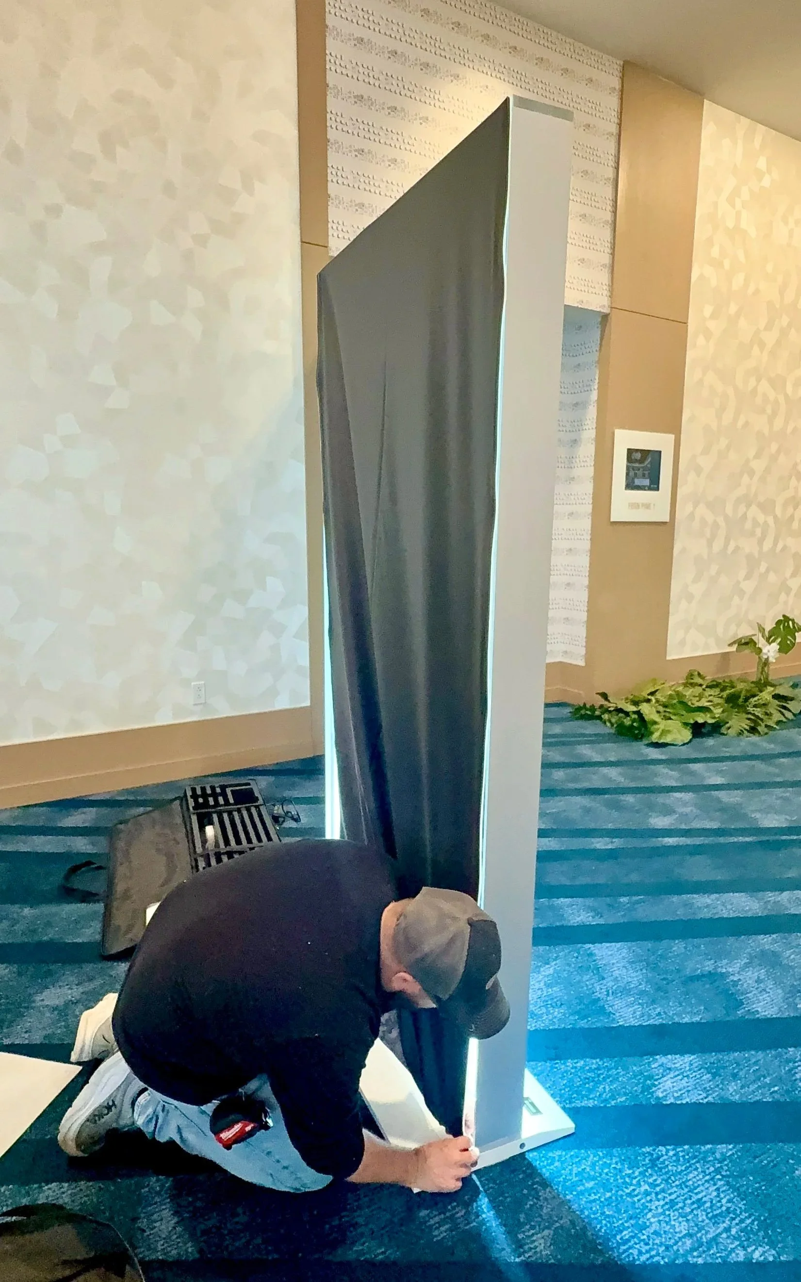 Jeremy, the installer, is wearing a black shirt, gray pants, and a gray cap kneels on the floor and screws a metal base to a freestanding, vertical, metallic panel in an indoor setting with patterned blue carpet and beige walls.