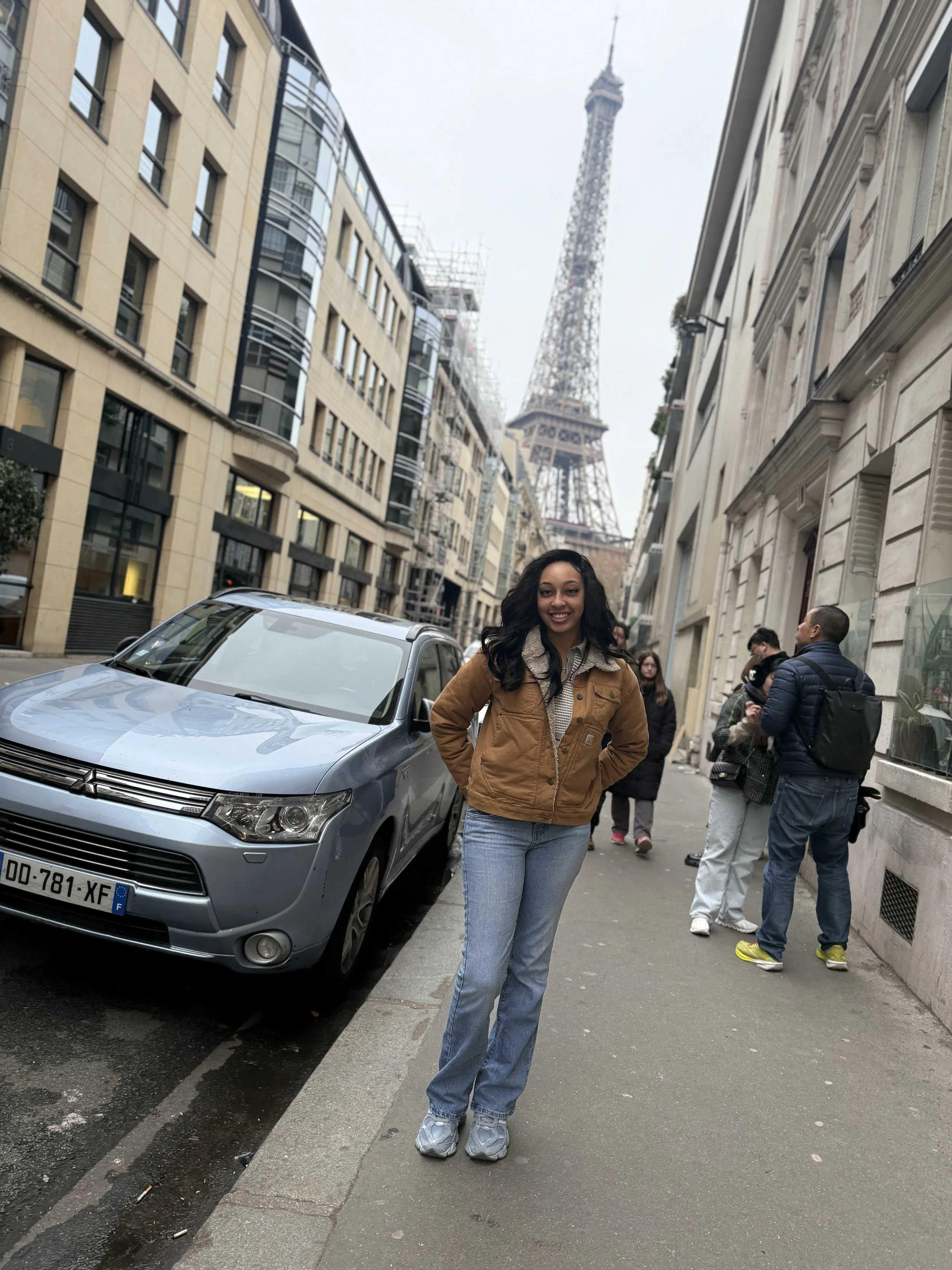 A woman with long black hair, wearing a brown jacket, blue jeans, and sneakers, standing on a sidewalk in Paris with the Eiffel Tower in the background.
