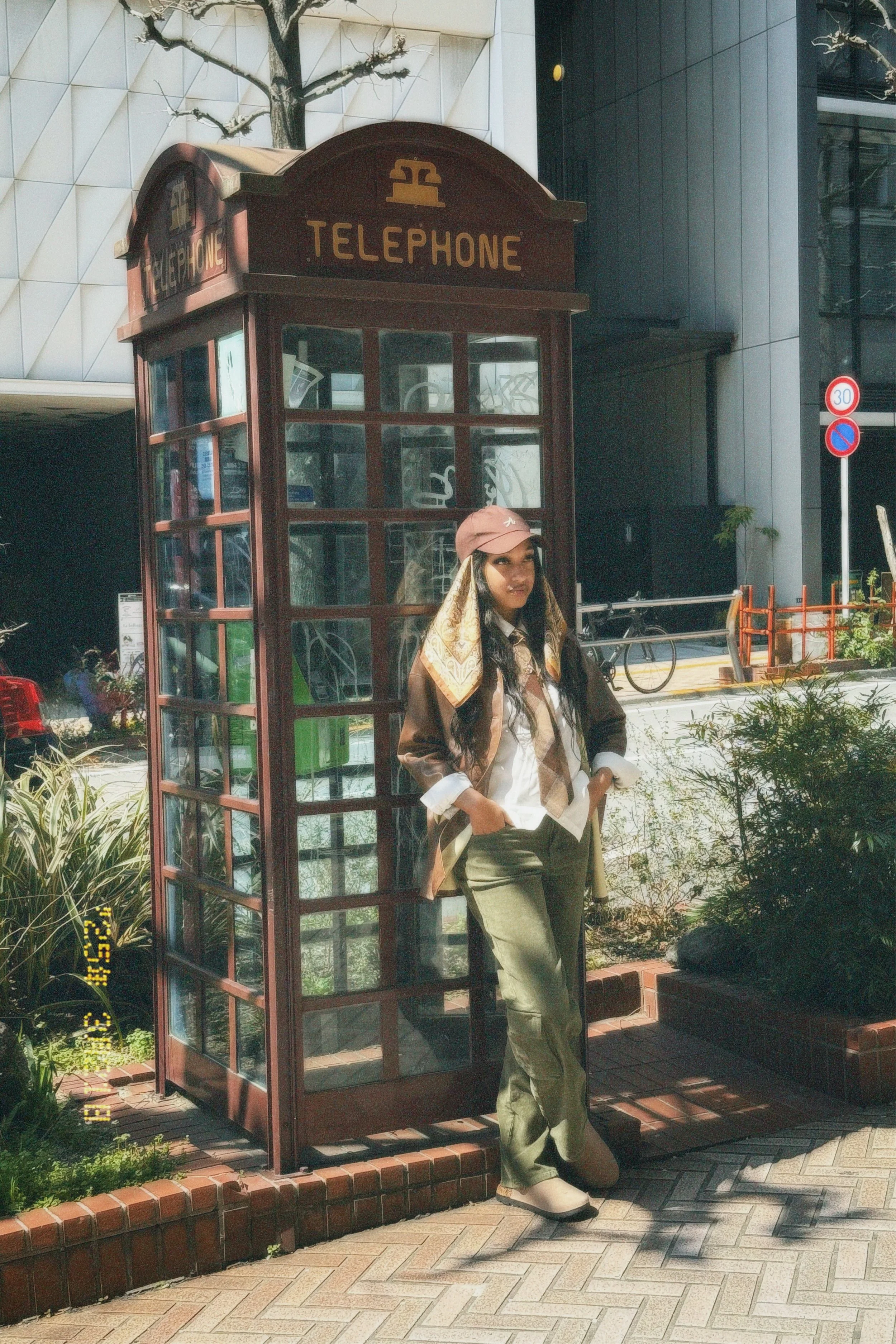 A young woman in casual clothing, wearing a pink cap, tan scarf, and khaki pants, standing next to a classic red telephone booth on a city sidewalk with trees and modern buildings in the background.