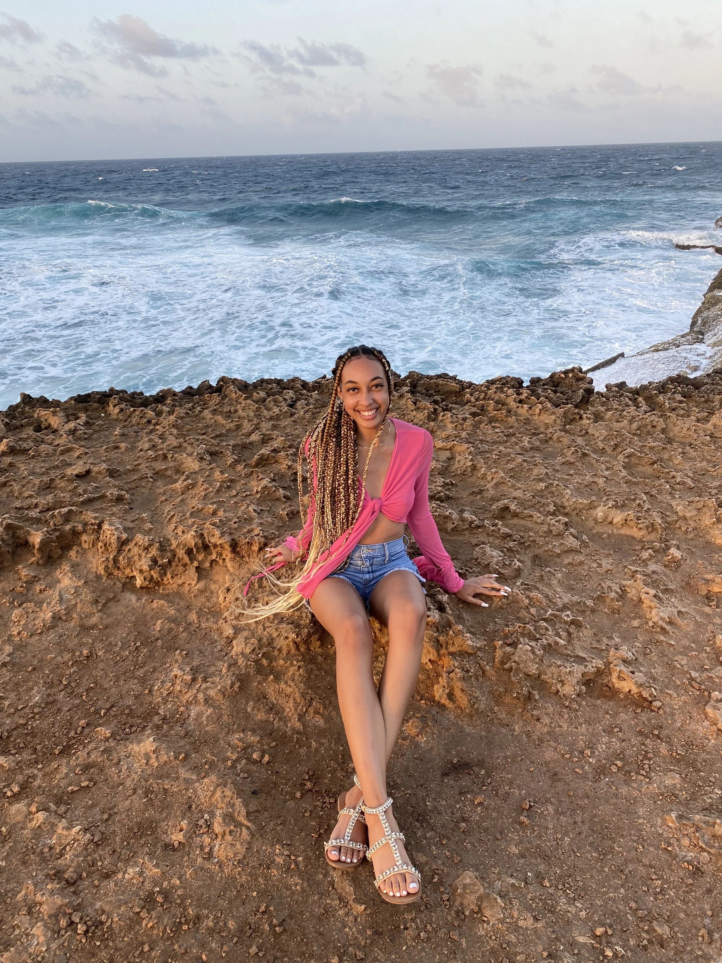 A young woman with braids sitting on rocky shoreline with ocean waves crashing behind her, wearing a pink top, denim shorts, and sandals, smiling at the camera.