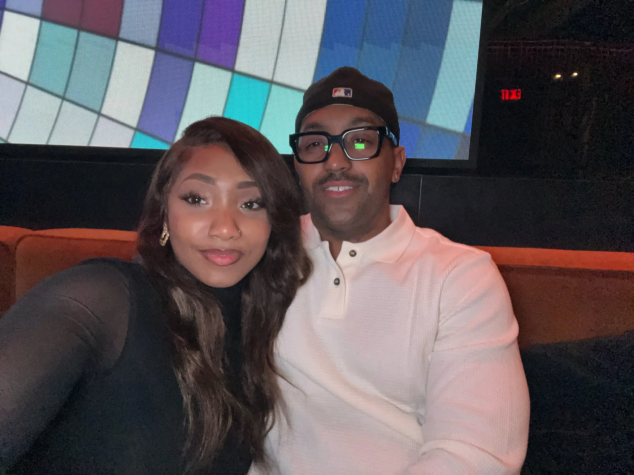 A couple sitting together in a dimly lit venue, with a colorful display screen behind them. The woman has long dark hair and is wearing a black top, and the man has glasses, a beard, a black baseball cap, and a white shirt.