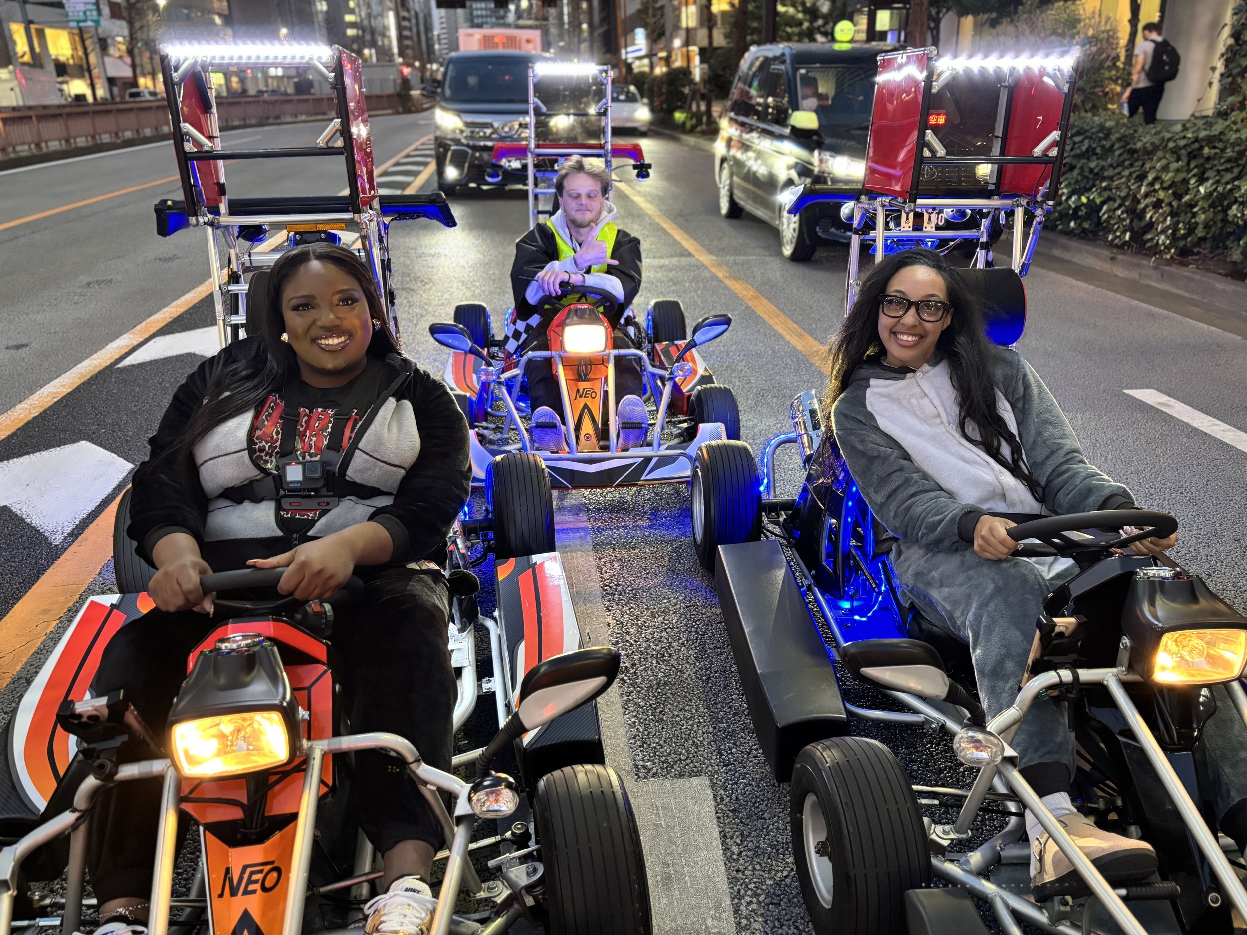 Three people sitting in go-karts on a city street at night, smiling. The street is wet, with cars in the background, and illuminated buildings and streetlights.
