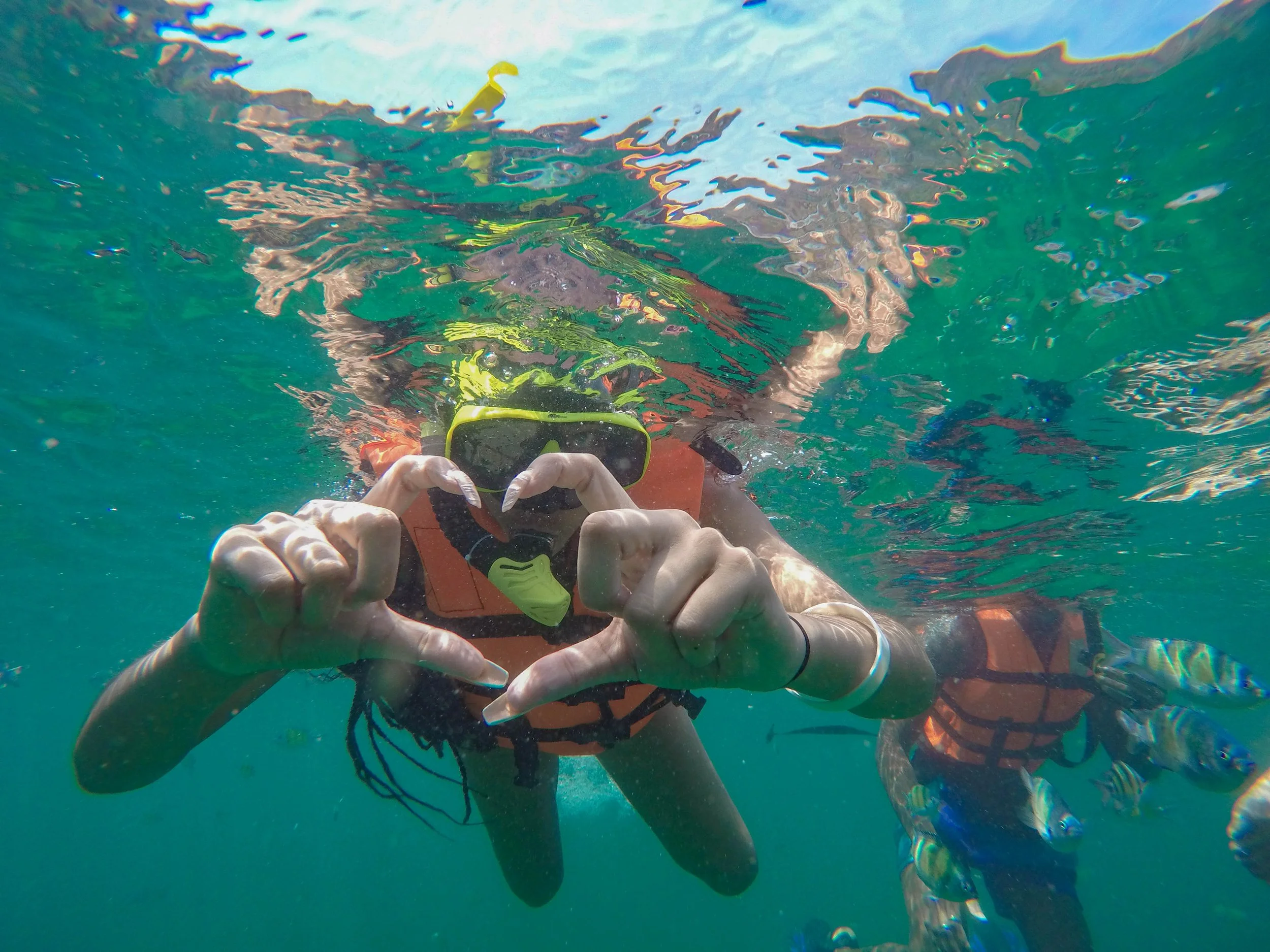 A person in a life jacket and snorkeling mask underwater making a heart shape with their hands.