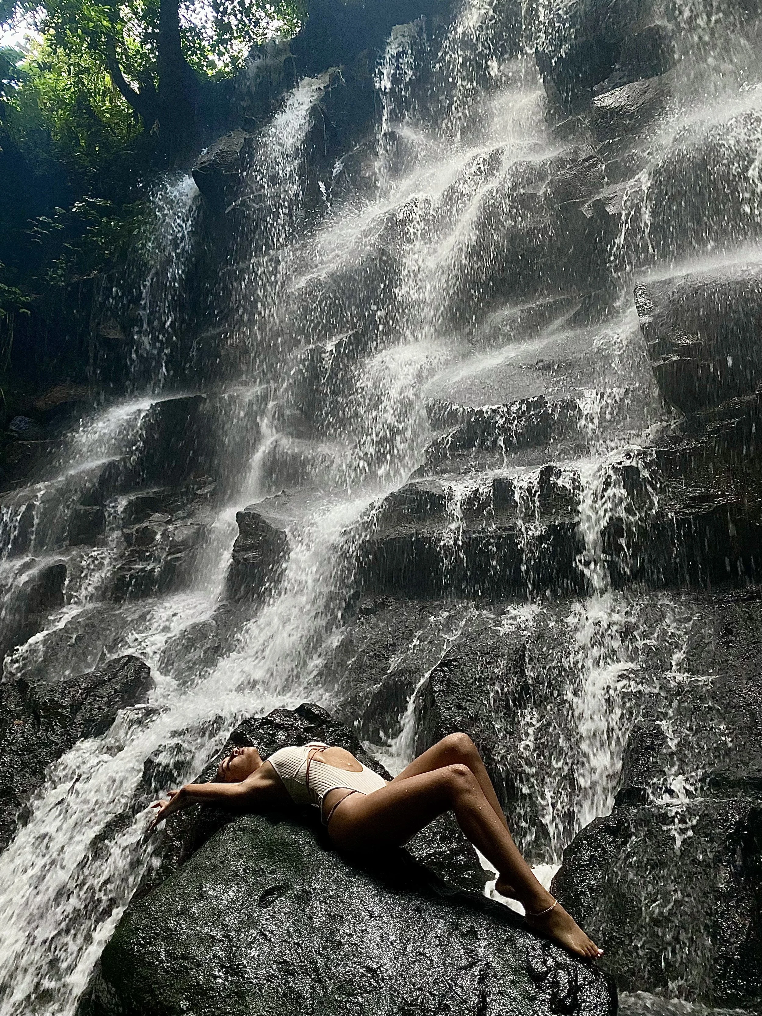 A woman in a swimsuit lying on a large rock under a waterfall in a lush, green forest.