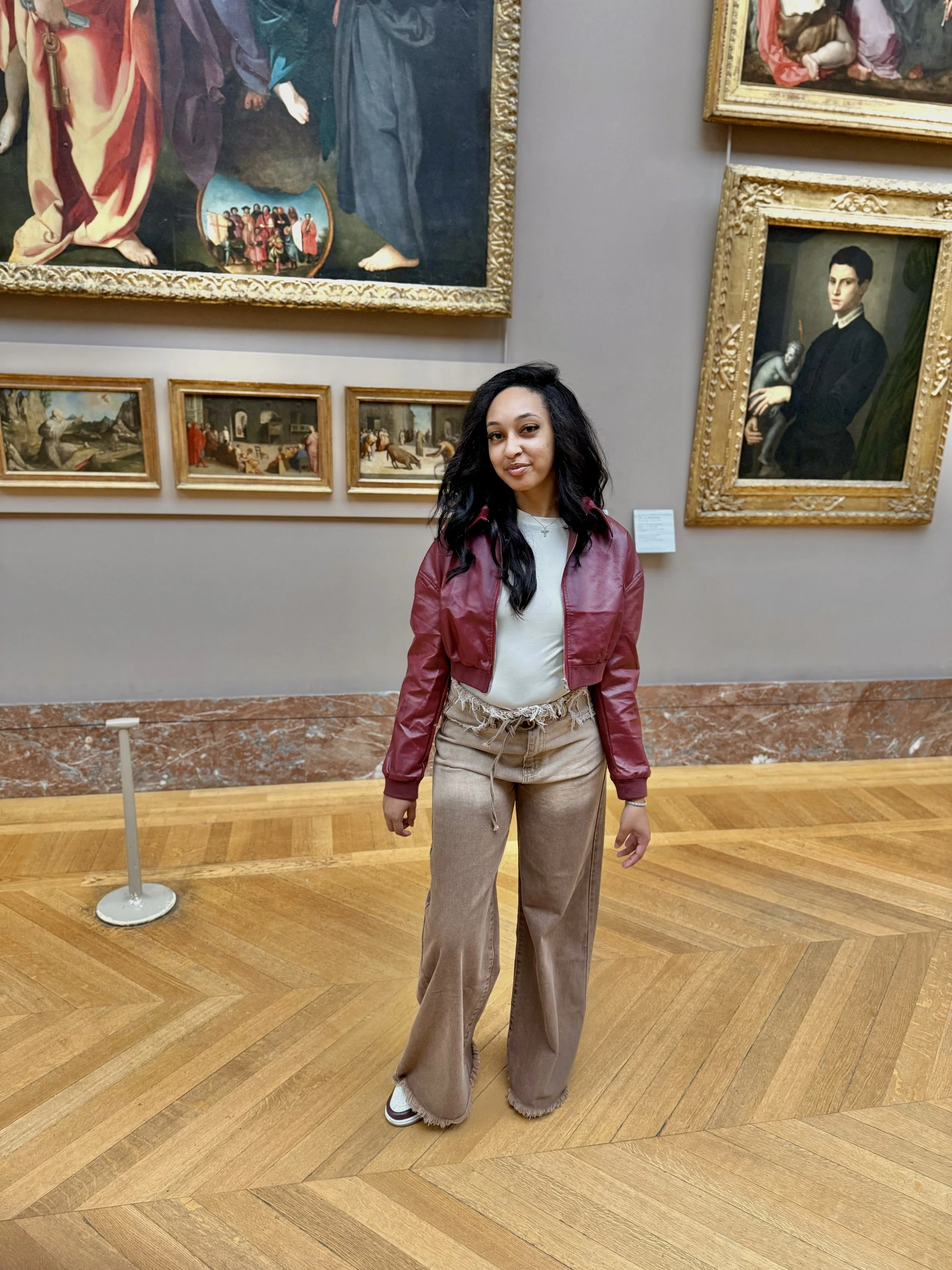 A woman with long black hair in a red leather jacket and beige wide-leg pants stands in an art gallery, surrounded by large framed paintings and smaller landscape artworks on the wall.