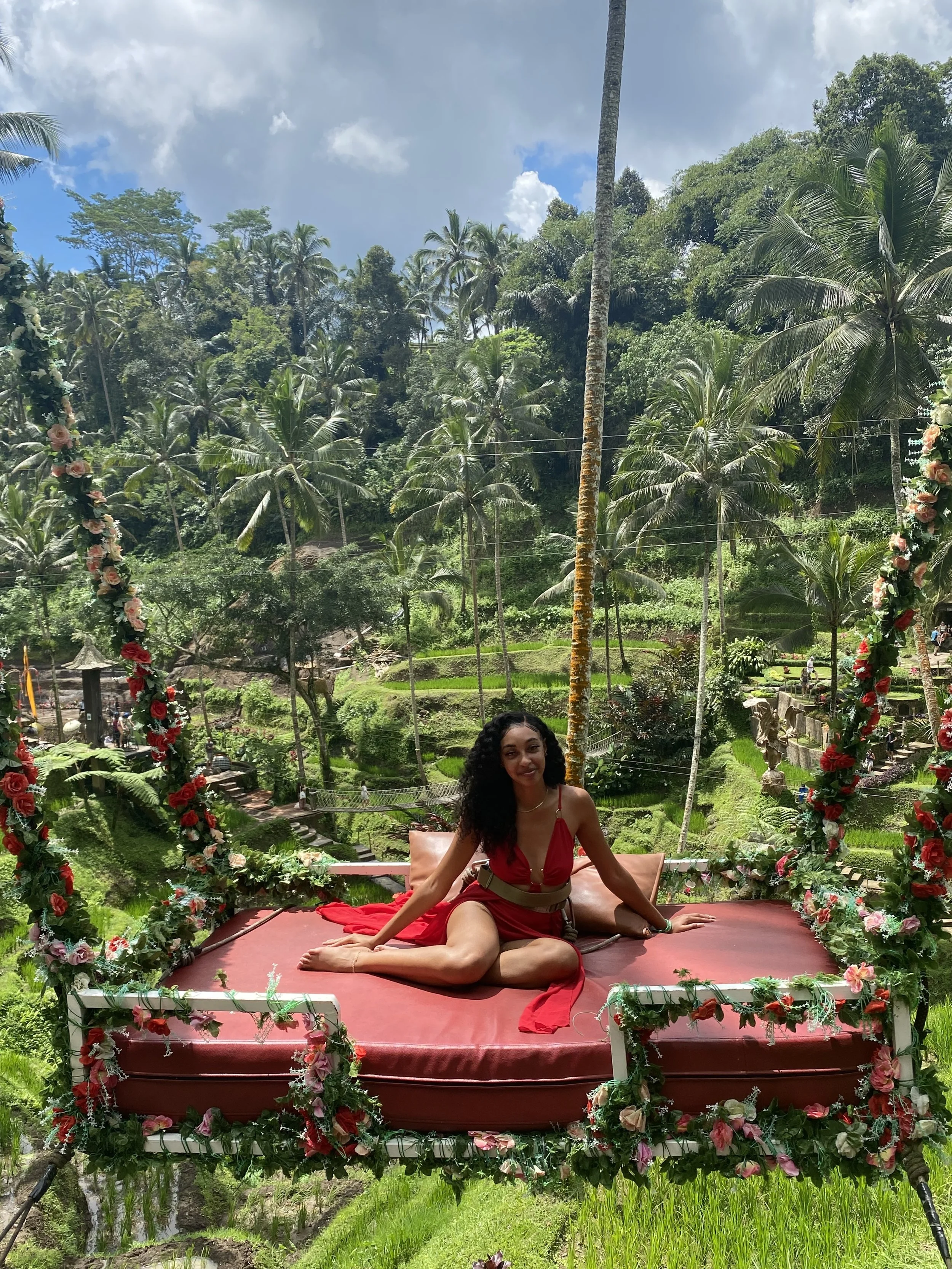 A woman in a red dress sitting on a decorated swing overlooking a lush tropical garden with palm trees.