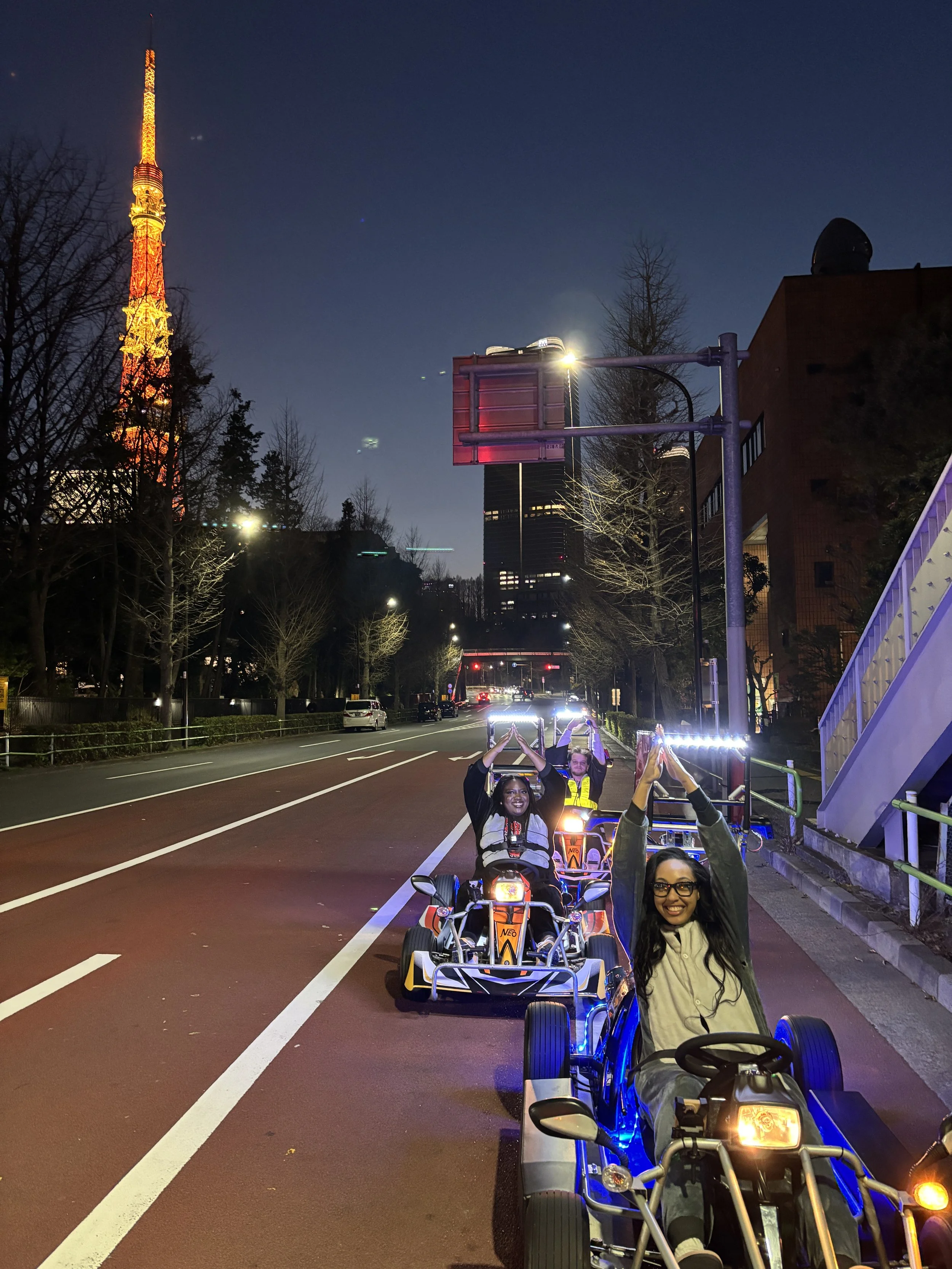 A group of four women riding go-karts at night on a city street, smiling and making heart shapes with their hands.