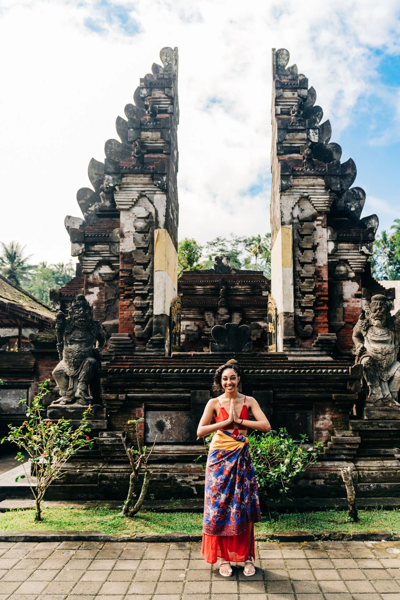 A smiling woman in a colorful traditional dress standing with hands folded in front of an ancient stone temple with intricate carvings and statues in Bali, Indonesia.