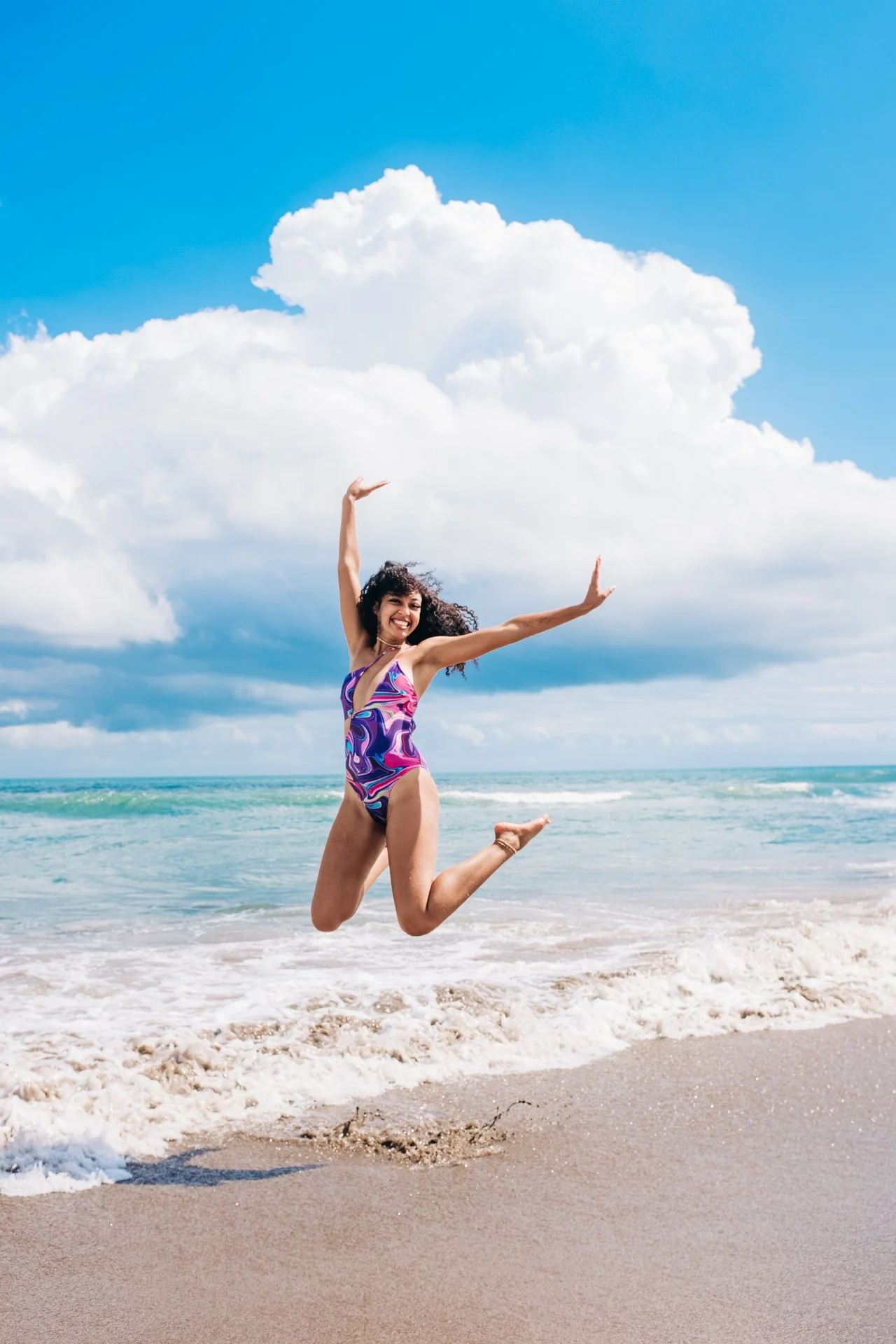 Woman jumping on the beach in a colorful swimsuit with clouds and blue sky in the background.