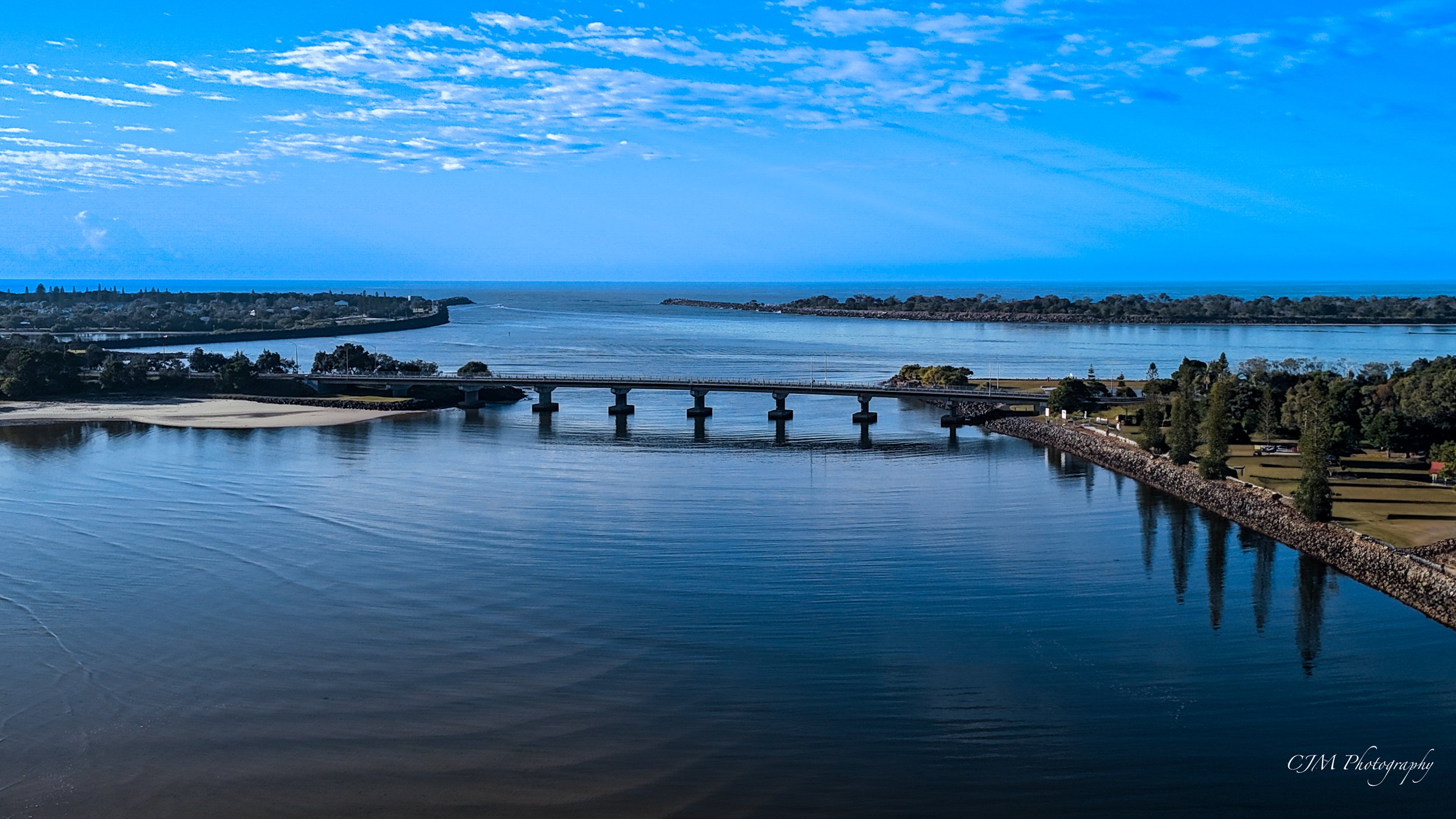 A scenic view of a river with a bridge, surrounded by trees and land, with a vast body of water and a blue sky with clouds in the background.
