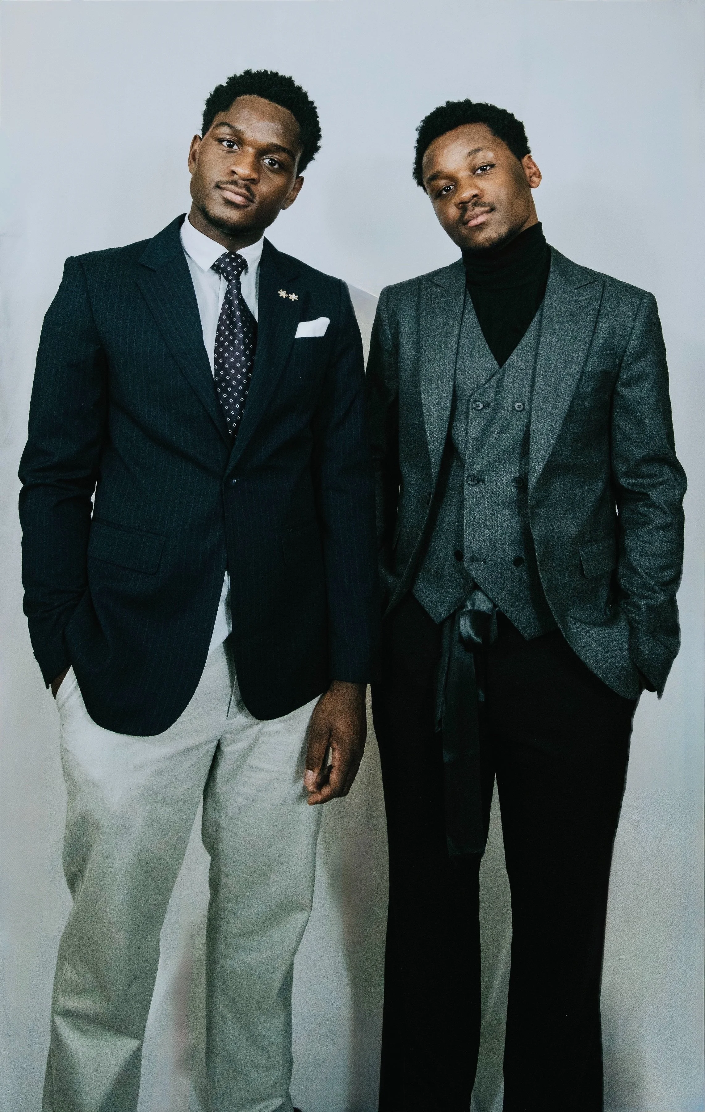 Two young men in formal suits standing together against a plain background, posing for the camera.
