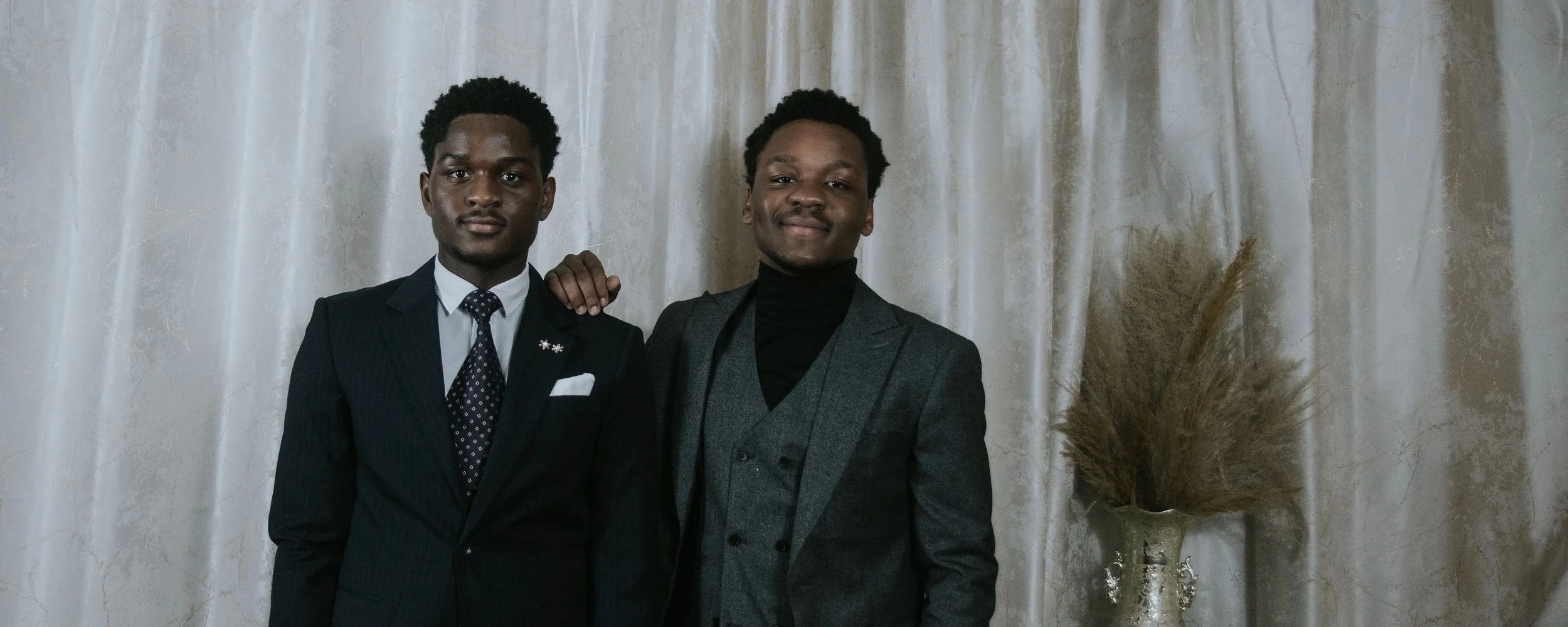 Two young men dressed in suits standing close together in front of a cream-colored curtain, with one man's hand resting on the other's shoulder, and a decorative vase with dried pampas grass next to them.