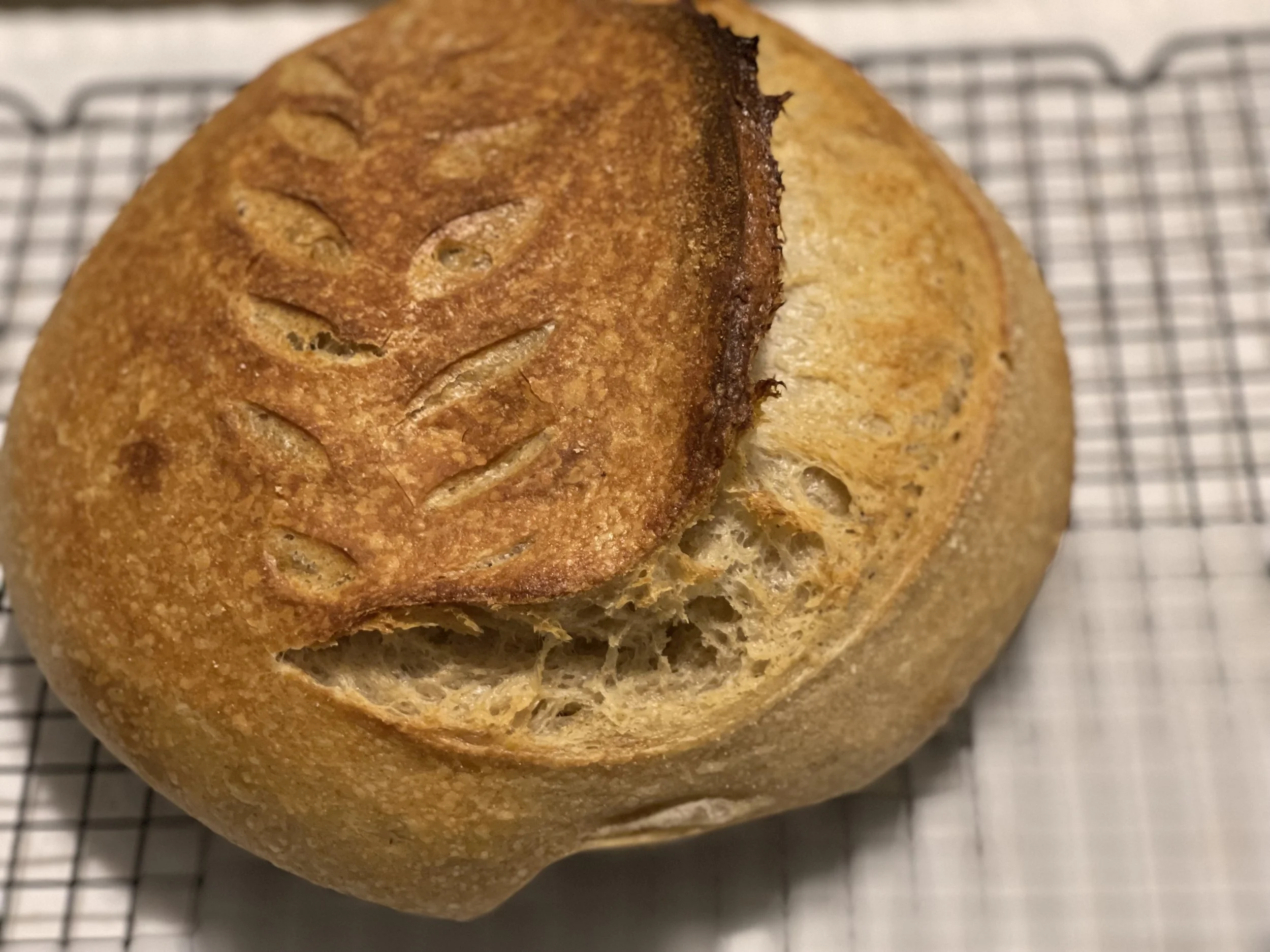 Close-up of a freshly baked round loaf of bread with a golden crust and decorative scoring on top, resting on a wire rack.