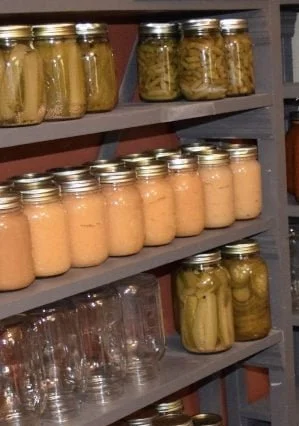 Multiple jars of preserved foods on a wooden shelf, including pickles and possibly canned fruits or vegetables.