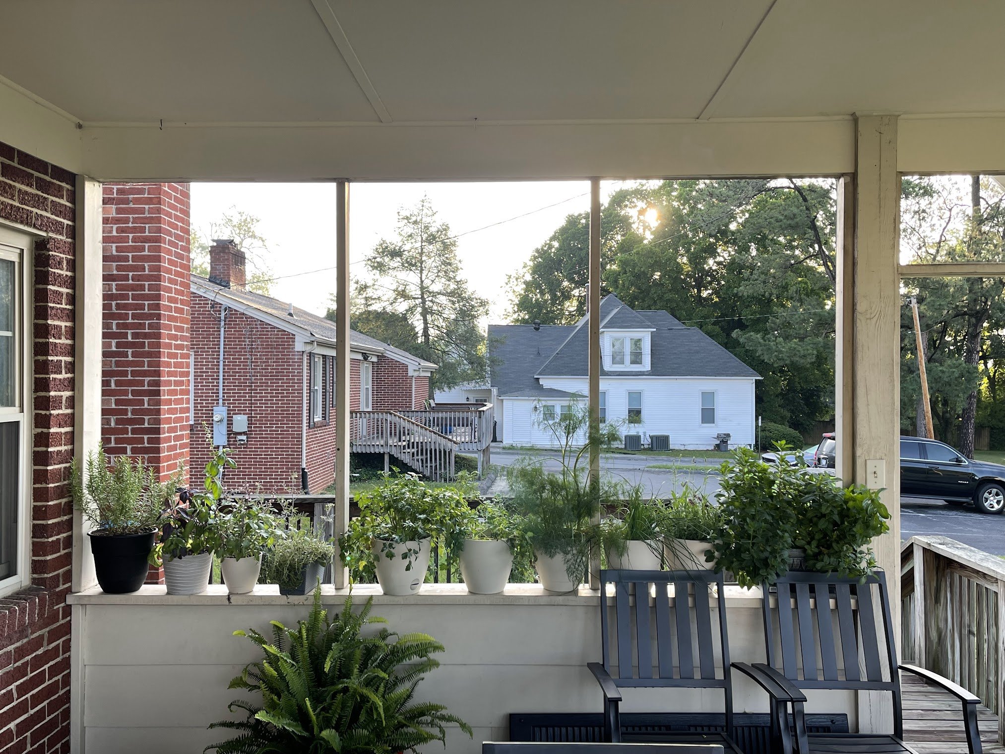 View from a porch with potted plants on the railing, showing neighboring houses, trees, and parked cars in a suburban neighborhood during daylight.